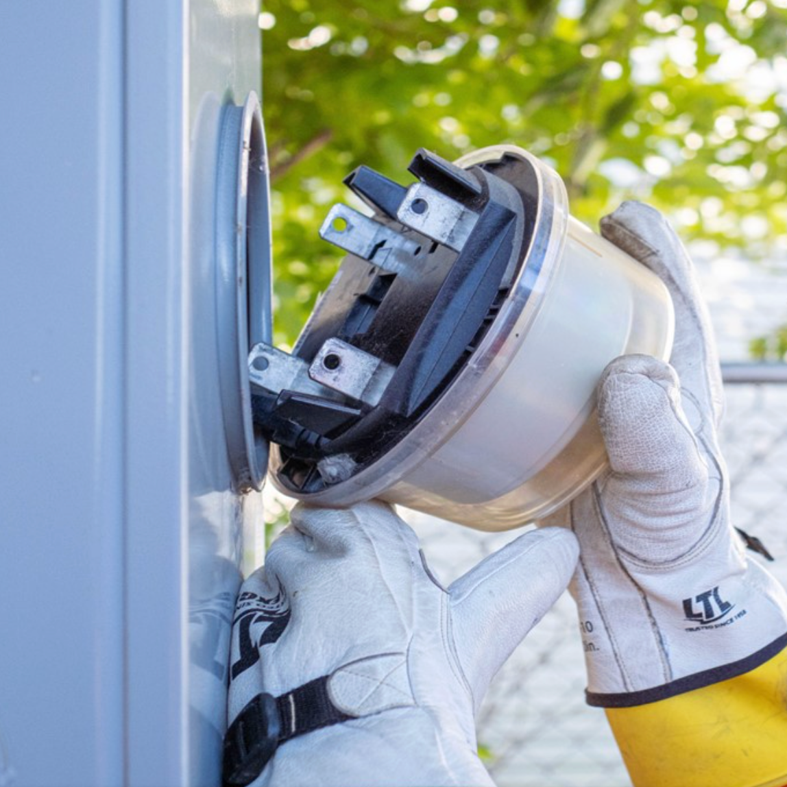 A person wearing white gloves is installing or repairing an electrical component inside an outdoor electrical box, with greenery in the background.