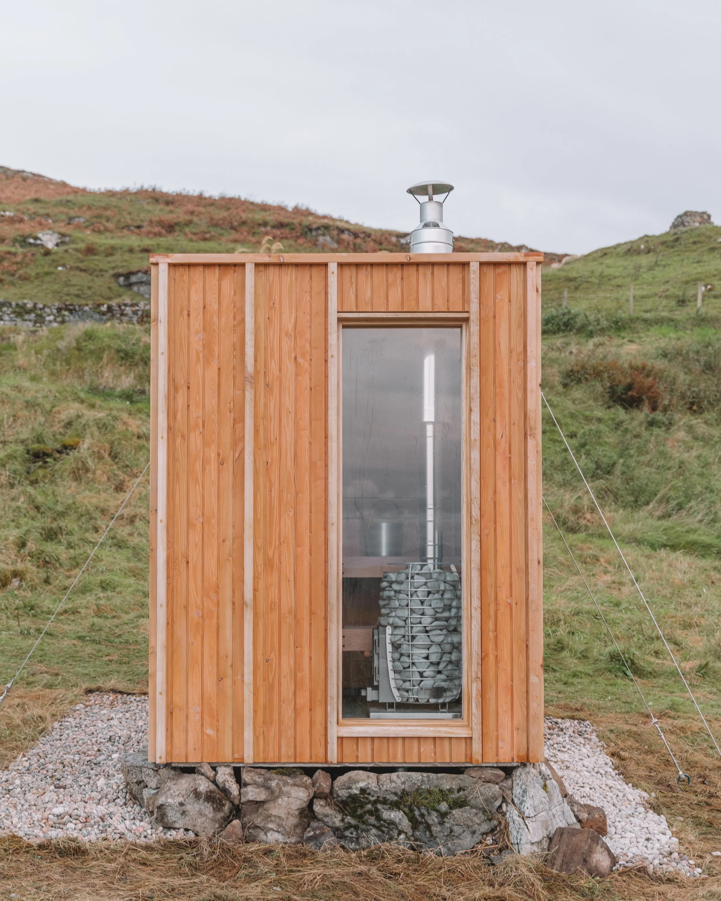Small wooden sauna with glass front door and rocks at its base, set in a grassy hillside landscape with cloudy sky.