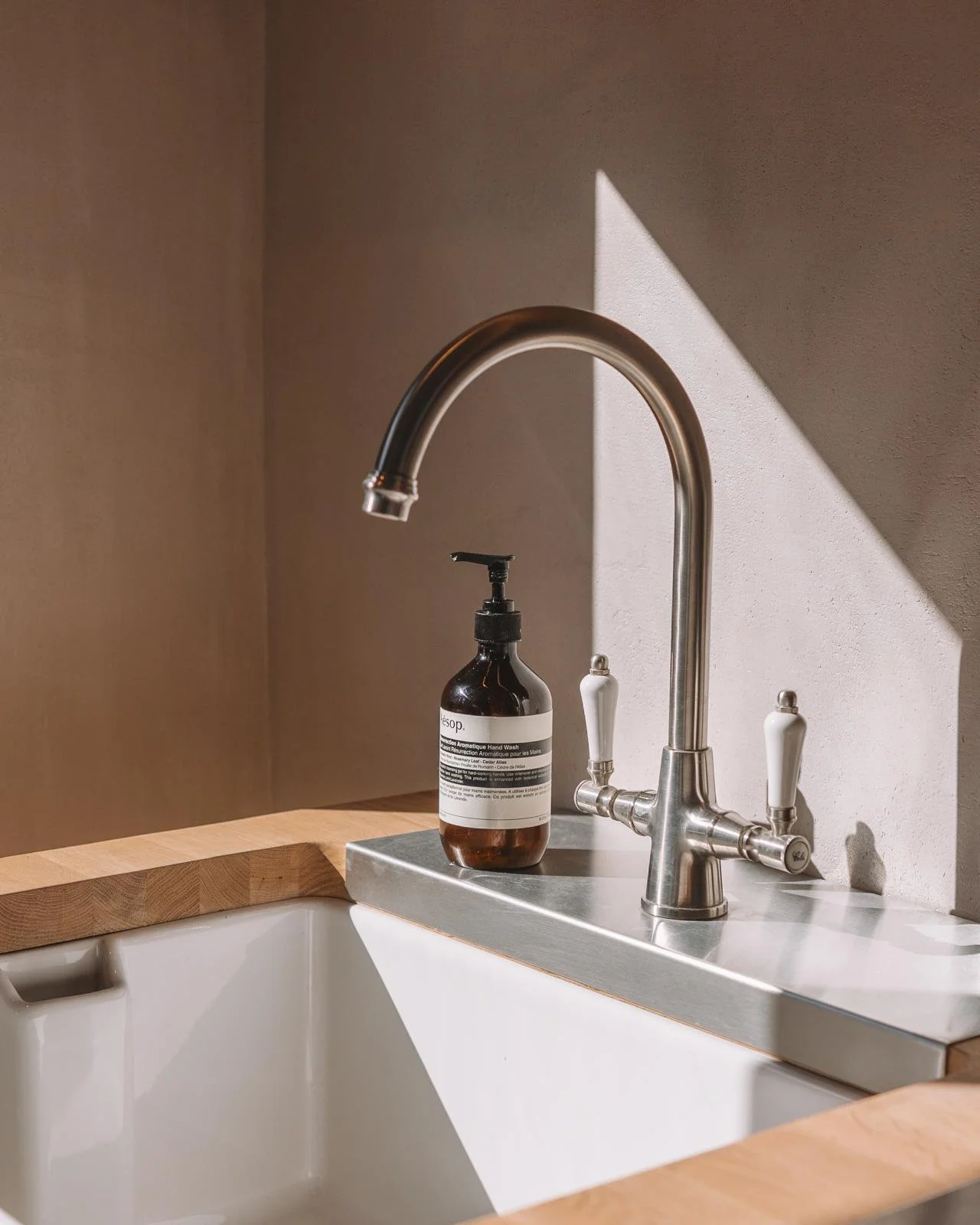 A kitchen sink with a modern silver faucet, a bottle of hand soap, and a wooden countertop. Sunlight creates a shadow on the wall behind the sink.