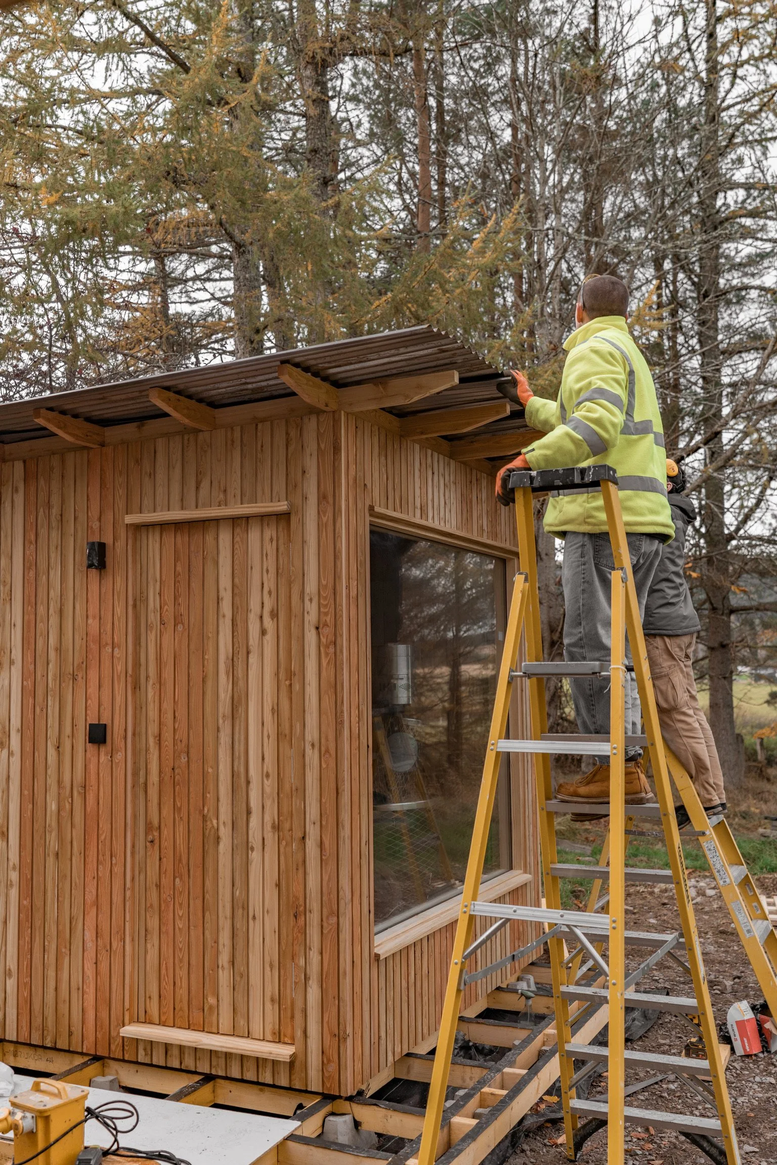 Two workers constructing or finishing a small wooden outdoor building, with a ladder, surrounded by trees.