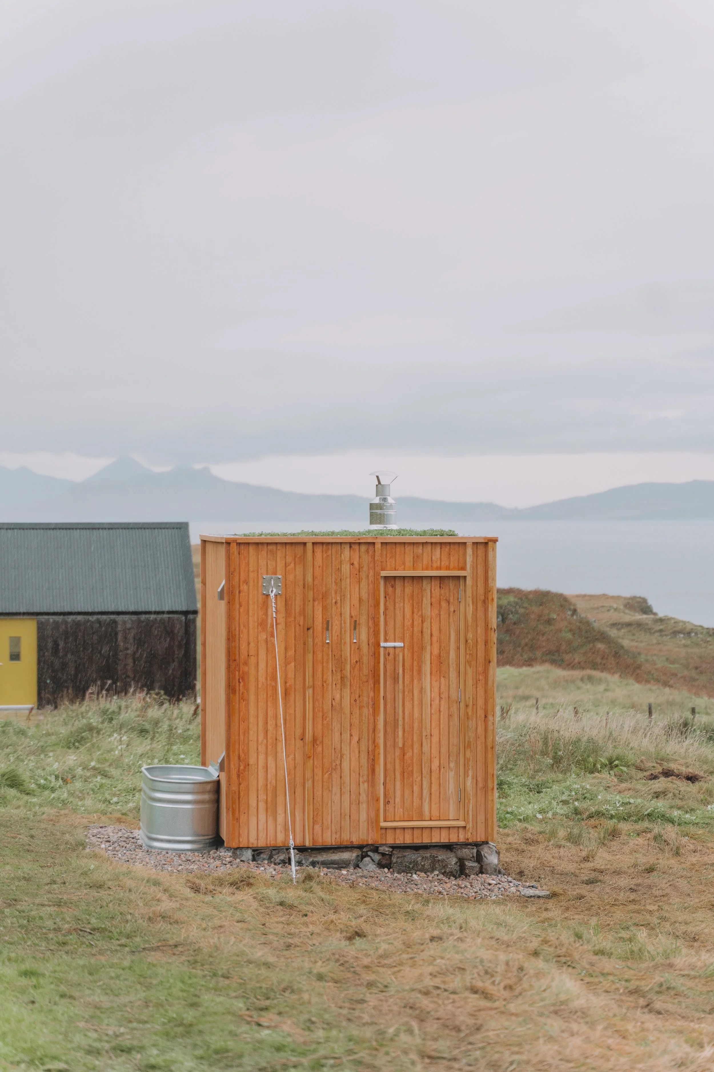 A small wooden outdoor sauna in a grassy landscape with a body of water and mountains in the background.