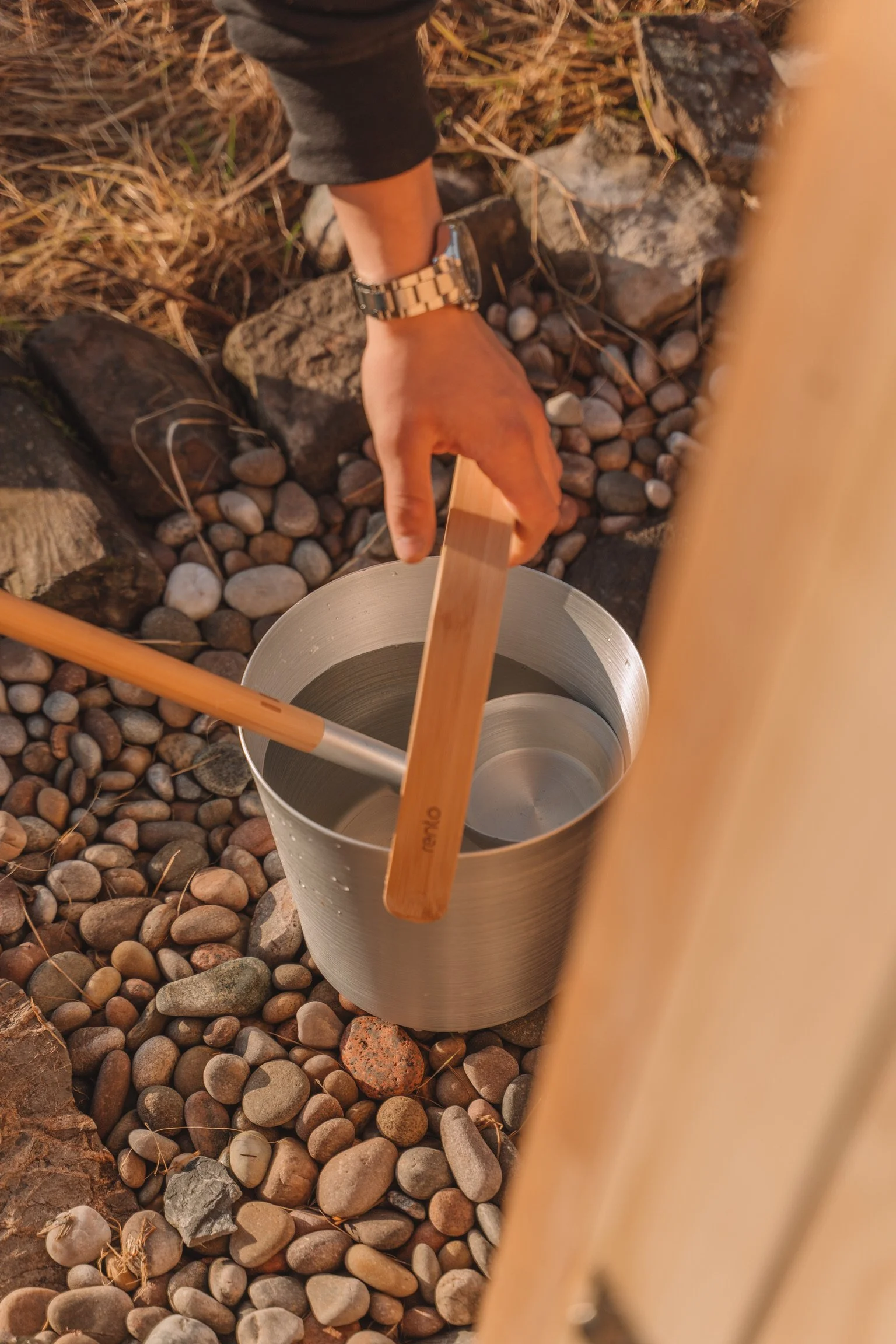 Close-up of a person pouring water into a stainless steel thermos with a wooden stirring stick, set on rocky ground.