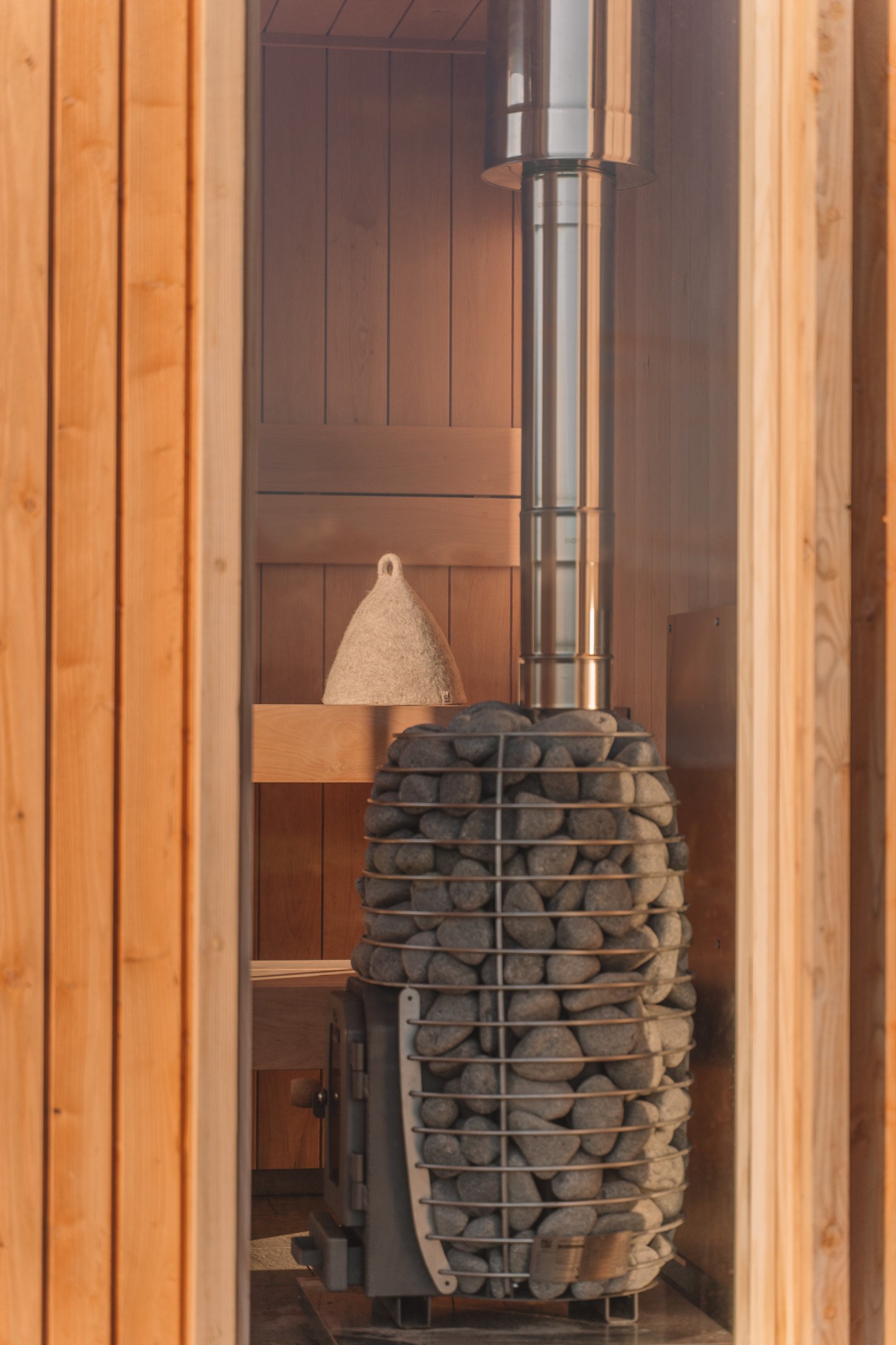 Interior view of a sauna with a wood-paneled wall, a metal chimney pipe, a heater filled with stones, and a wooden bench with a felt hat on top.