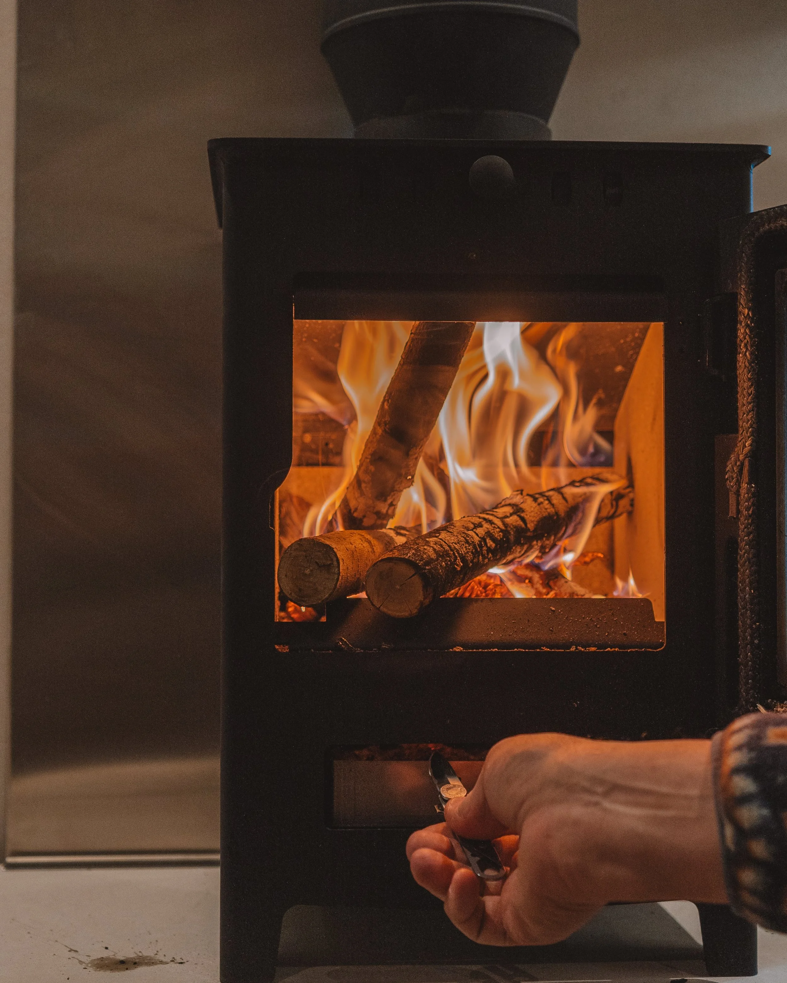 A person using tongs to add wood logs into a small black wood-burning stove with flames inside.