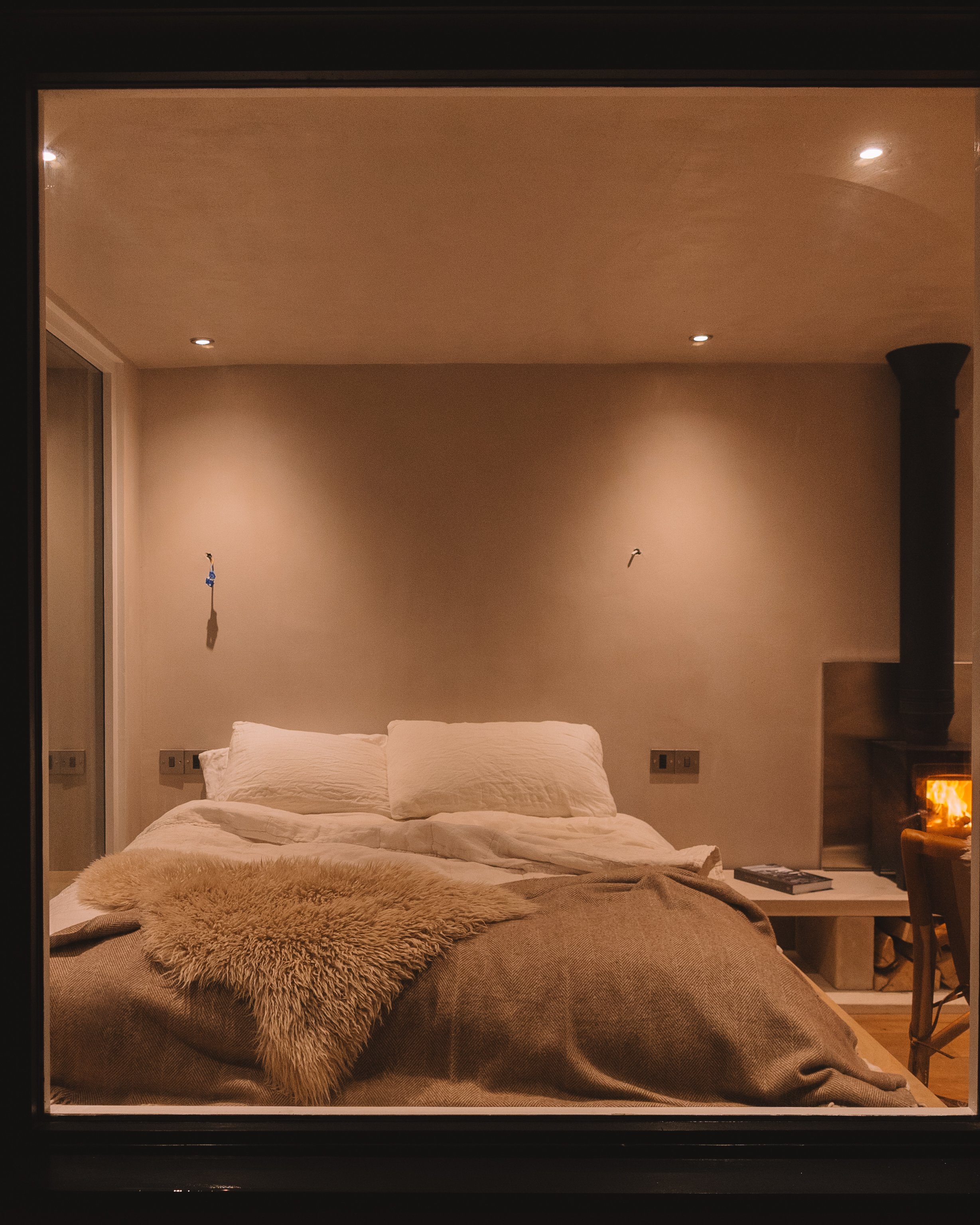 A cozy bedroom scene with a bed covered in beige bedding and pillows, a fur throw blanket, a small side table, a book, and a lit fireplace, viewed through a window at night.