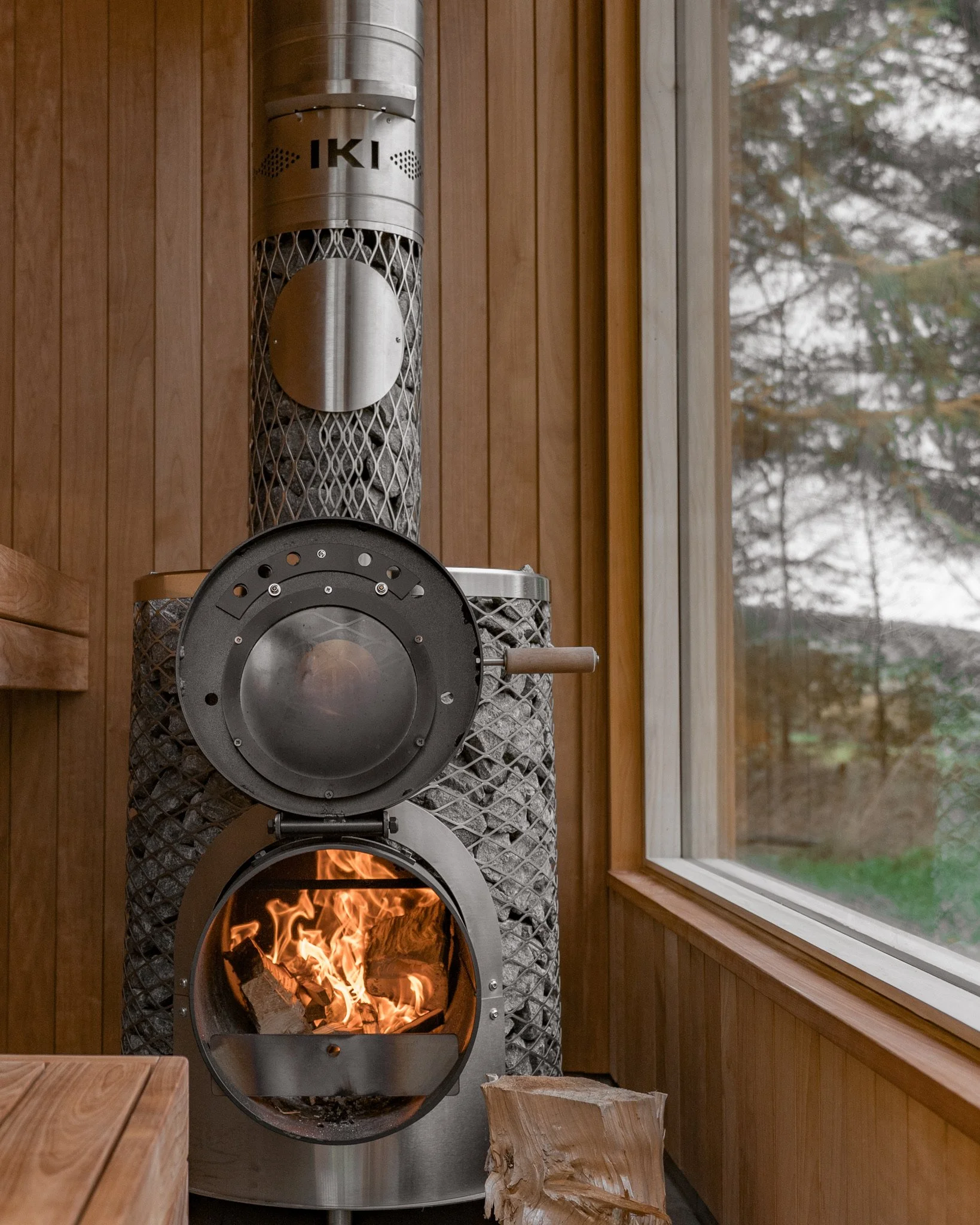 A modern wood-burning stove with visible flames, located indoors next to a window with a view of trees outside. The stove is made of metal and has a cylindrical shape, with a circular front door that is open, revealing the fire inside. The stove has a tall, chimney pipe extending upward.