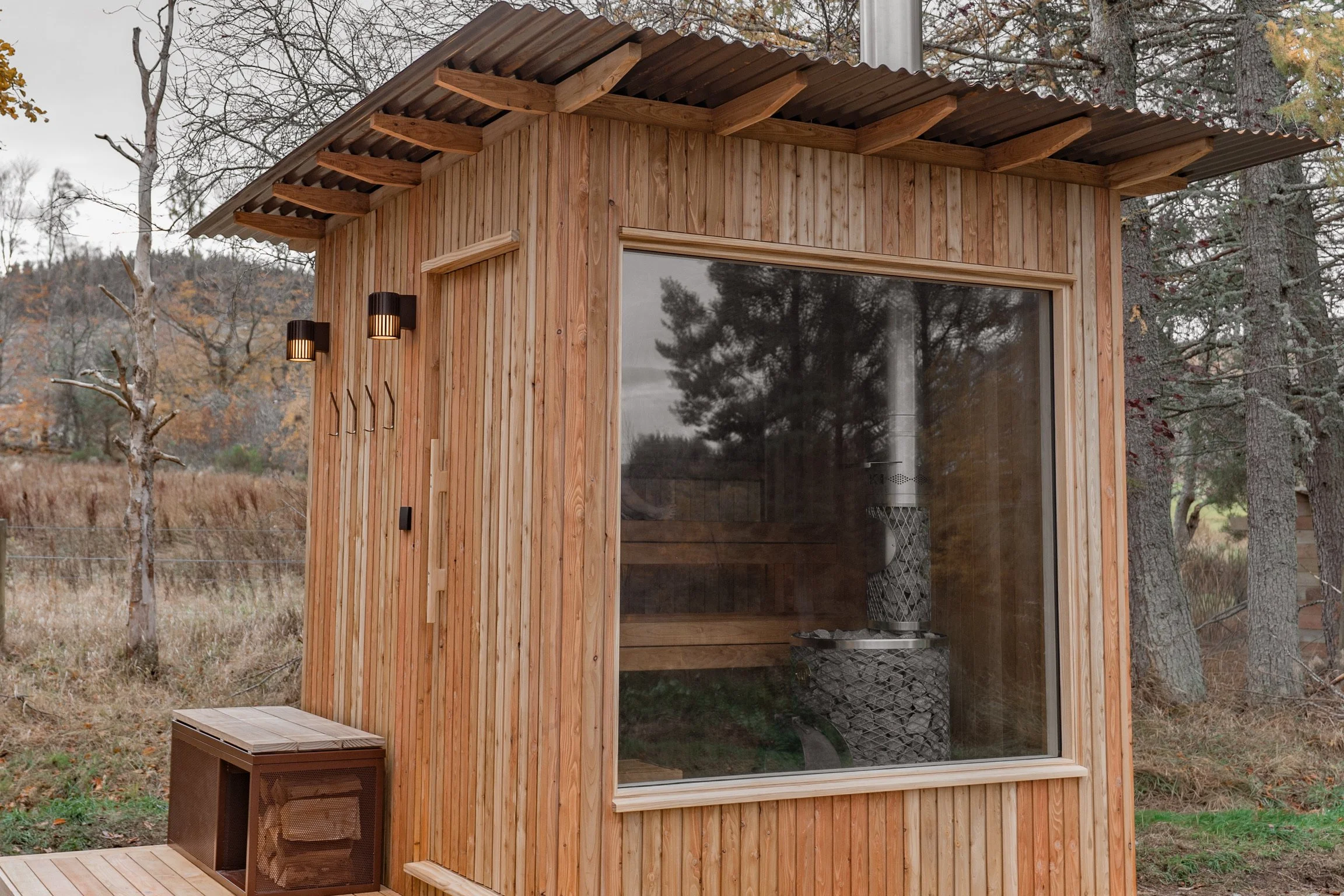 Small wooden cabin with large window, outdoor lights, and metal stove inside, set in a forested area.