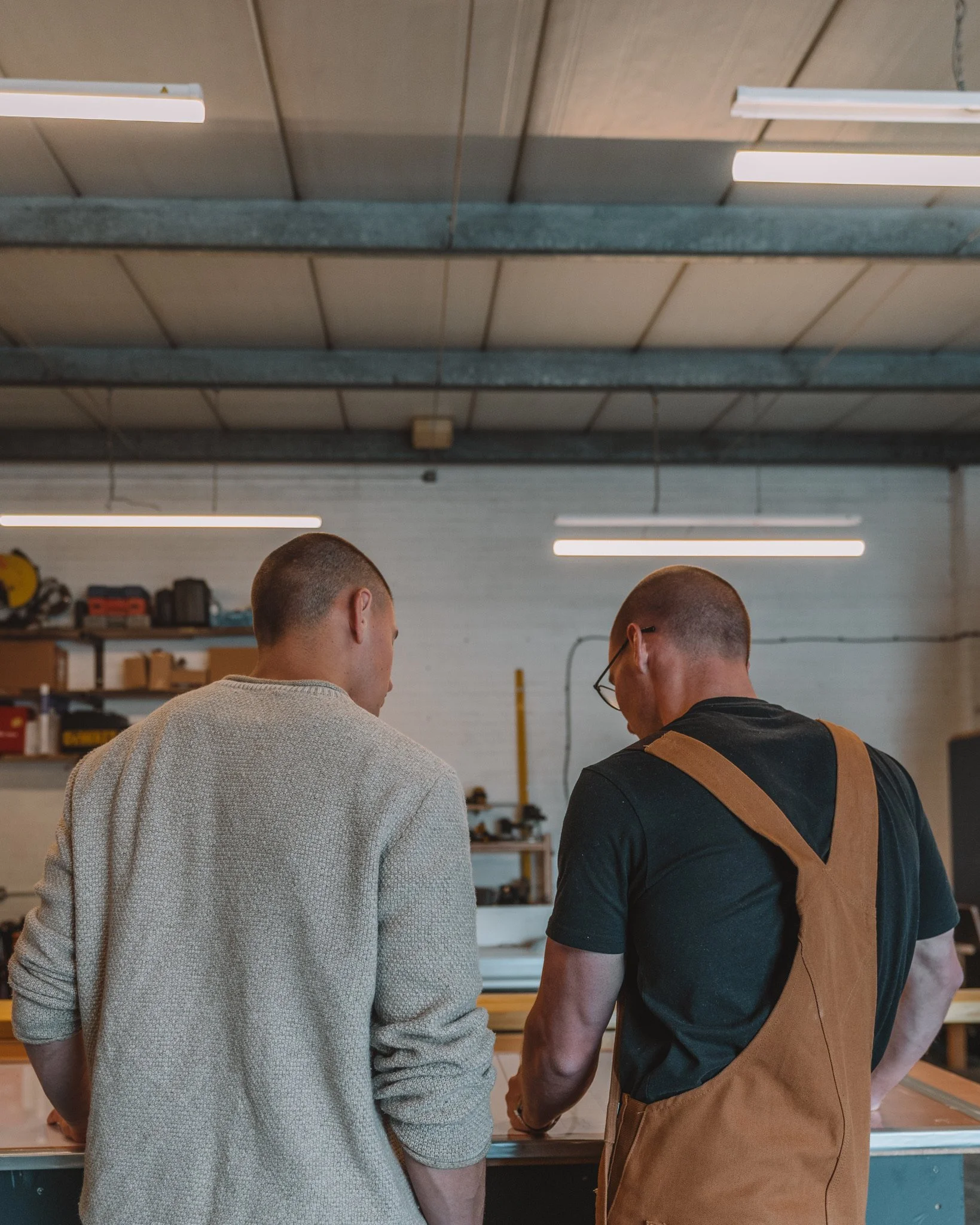 Two men are standing in a workshop, looking down at a worktable, with shelves and tools in the background.