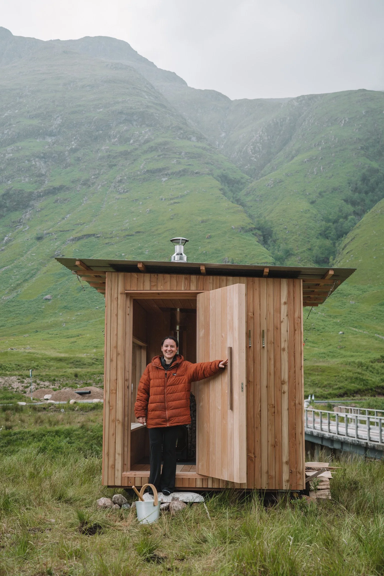 A woman in an orange jacket standing outside a small wooden cabin in a lush green valley with mountains in the background, smiling with her hand on the open door.