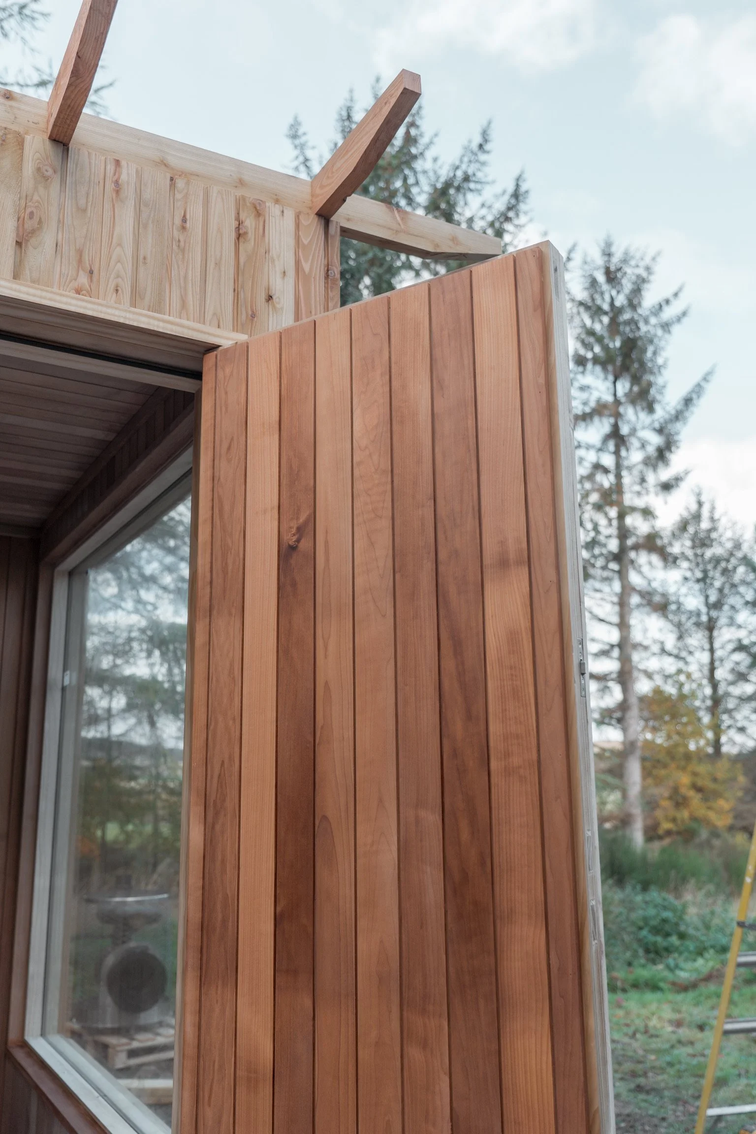 Wooden exterior wall of a building under construction with visible wooden siding, a window, and an open doorway. Part of a ladder and outdoor trees are visible in the background.
