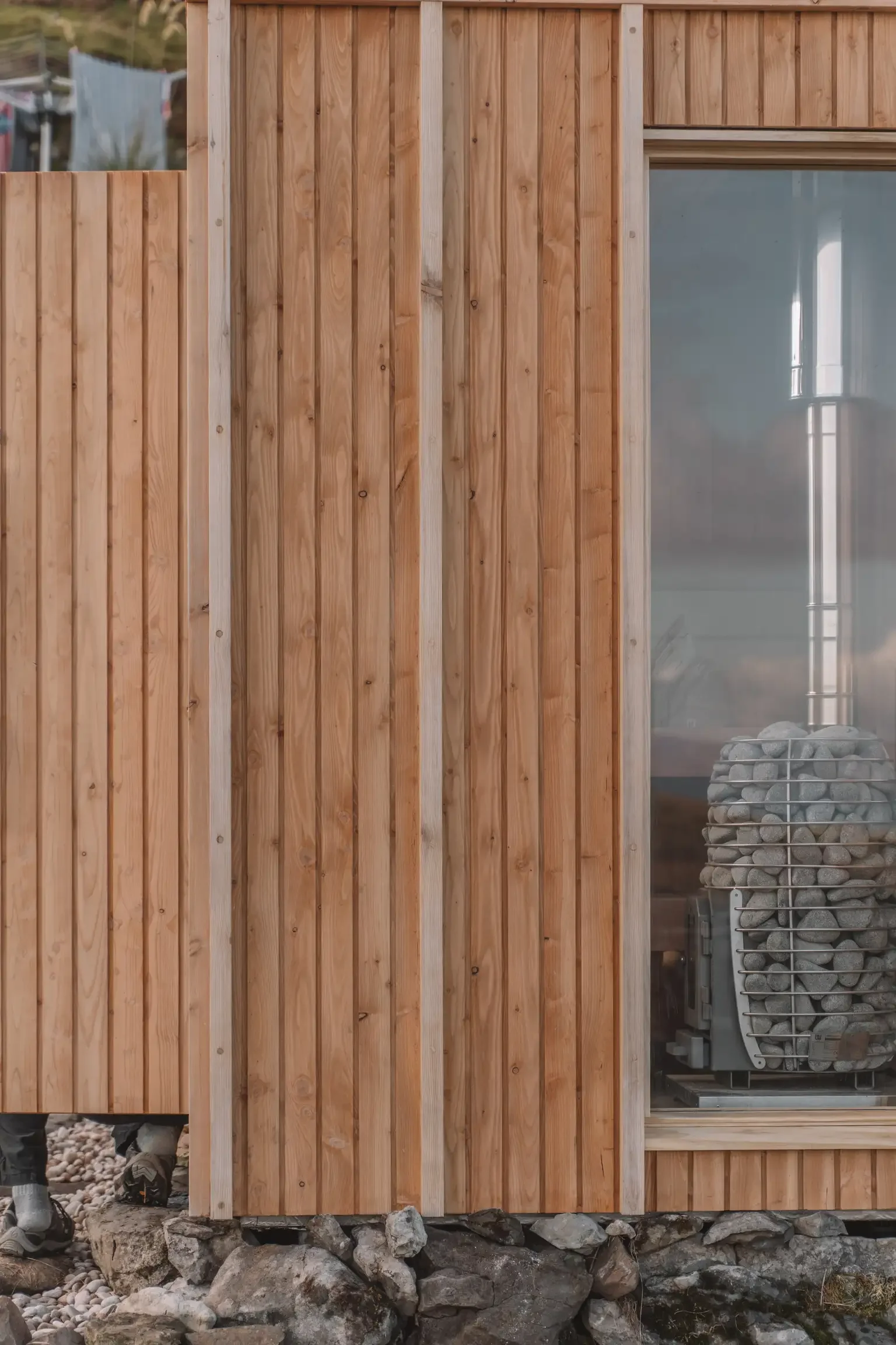 Close-up of a wooden exterior wall of a building, showing a glass window with a stack of rocks behind it, and some pebbles and rocks on the ground at the base.