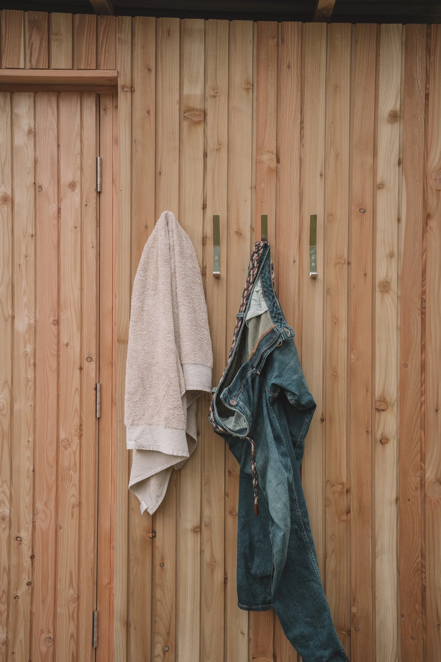 A wooden wall with three metal hooks, one holding a beige towel and the other a pair of blue denim jeans with a striped belt.
