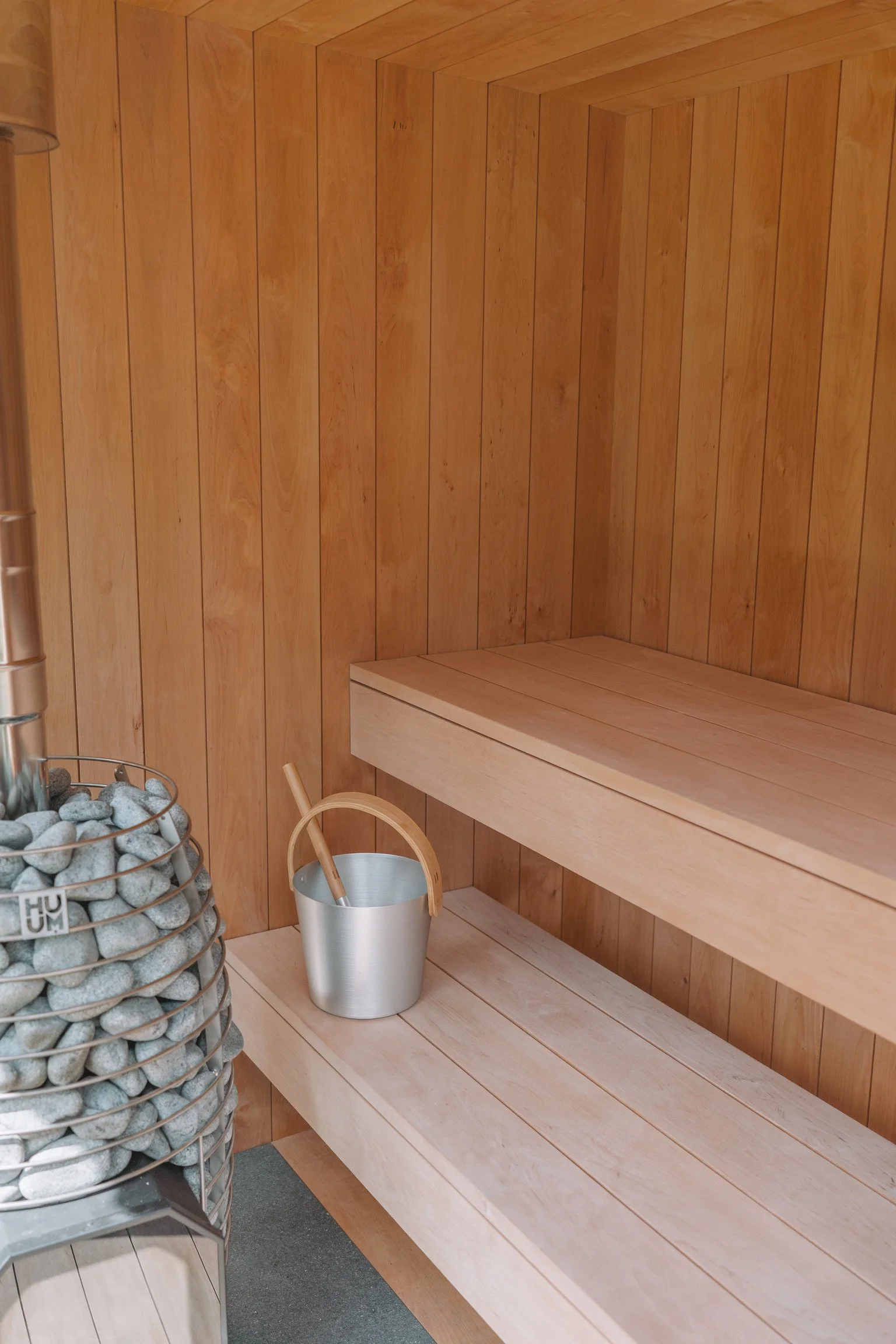 A wooden sauna interior with horizontal wood paneling, a wooden bench, and a metal bucket with a wooden ladle. Partially visible on the left is a container filled with sauna stones.