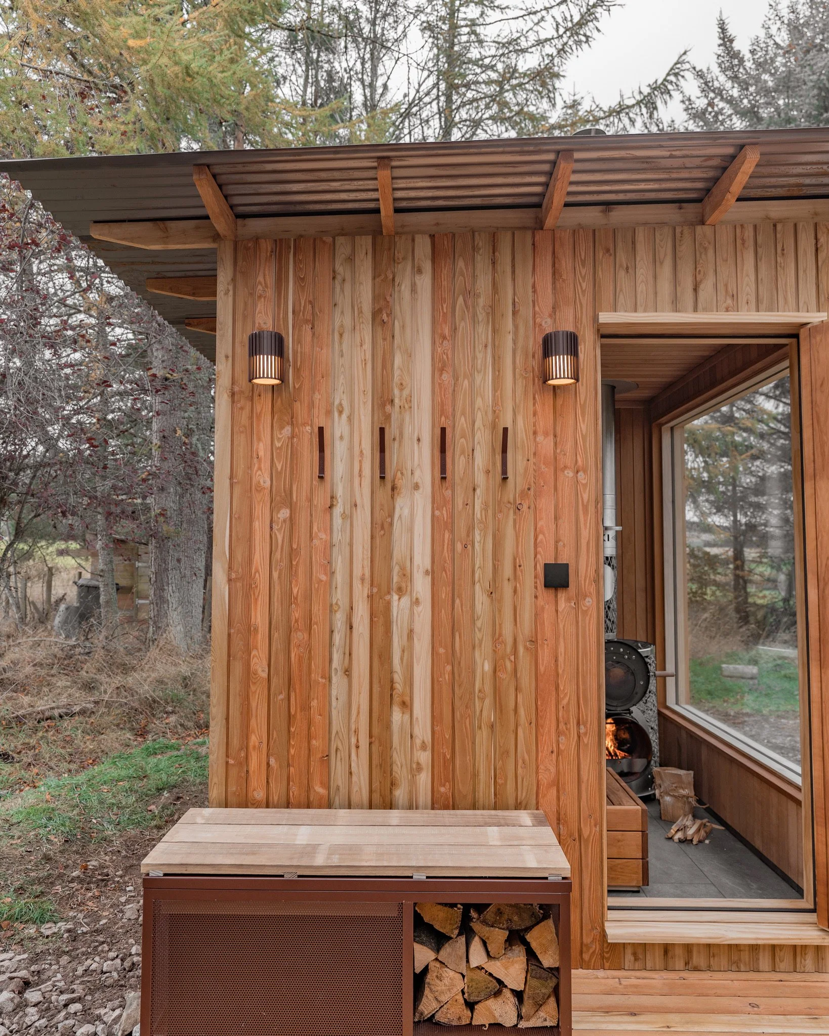 Exterior view of a tiny wooden cabin with vertical wood panels, black wall-mounted lights, and an open door revealing a wood stove and logs inside. A wood storage box is beneath a bench outside. Trees are visible in the background.