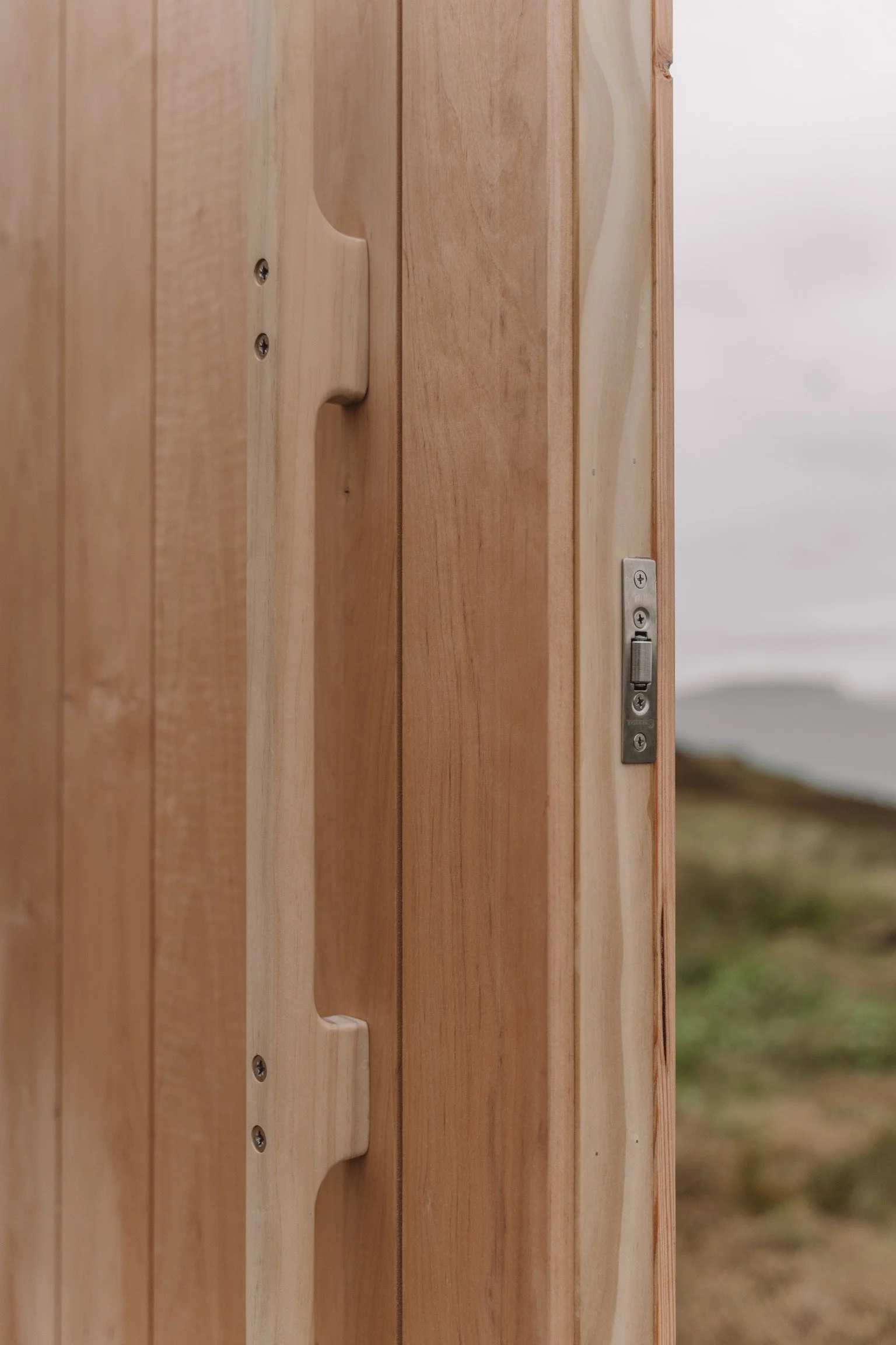 Close-up of a wooden door latch and lock on a natural wood door with an outdoor landscape background.