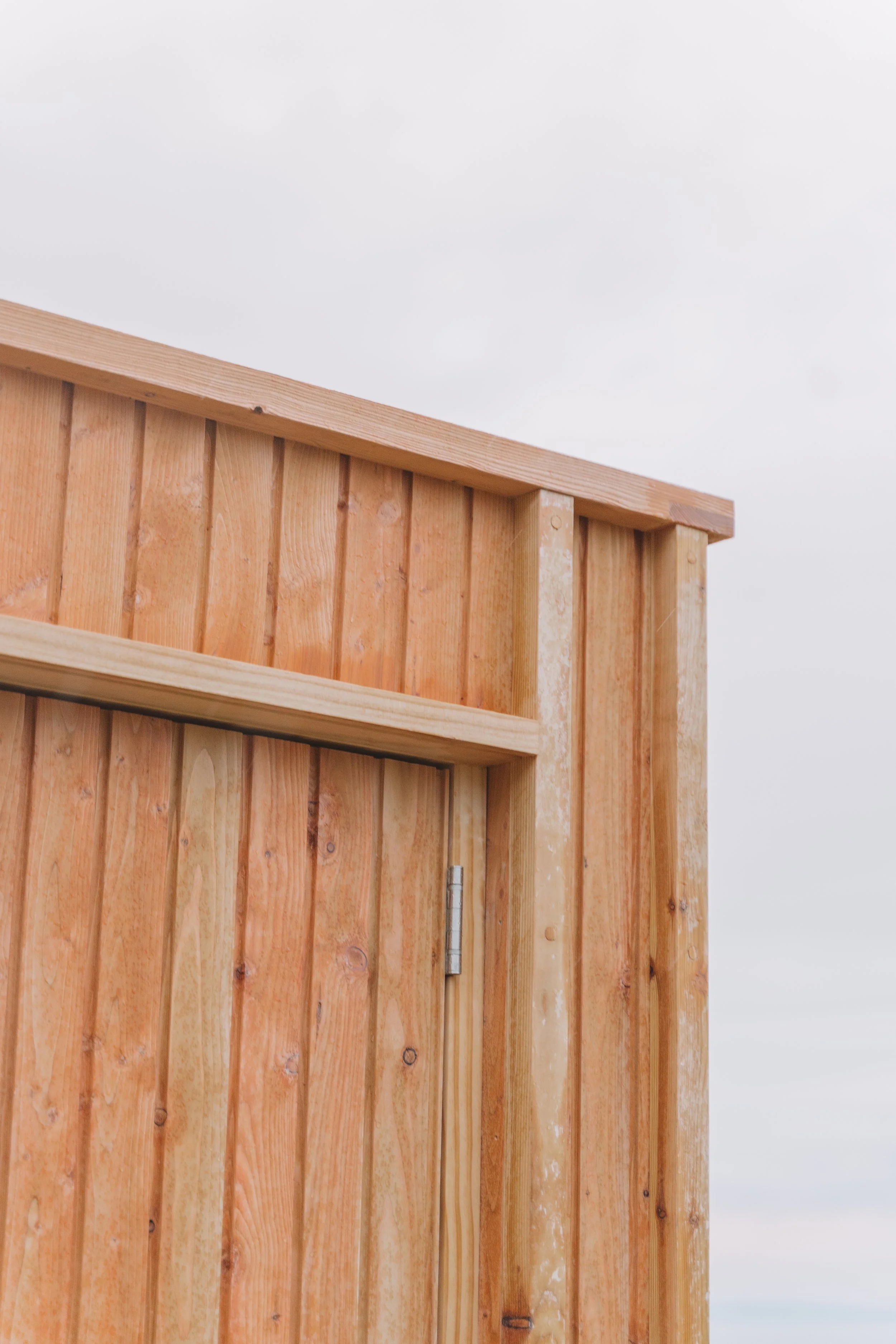 Close-up of a wooden structure, possibly a door or wall, made of vertical wooden planks with a hinge visible on the side, against a cloudy sky.