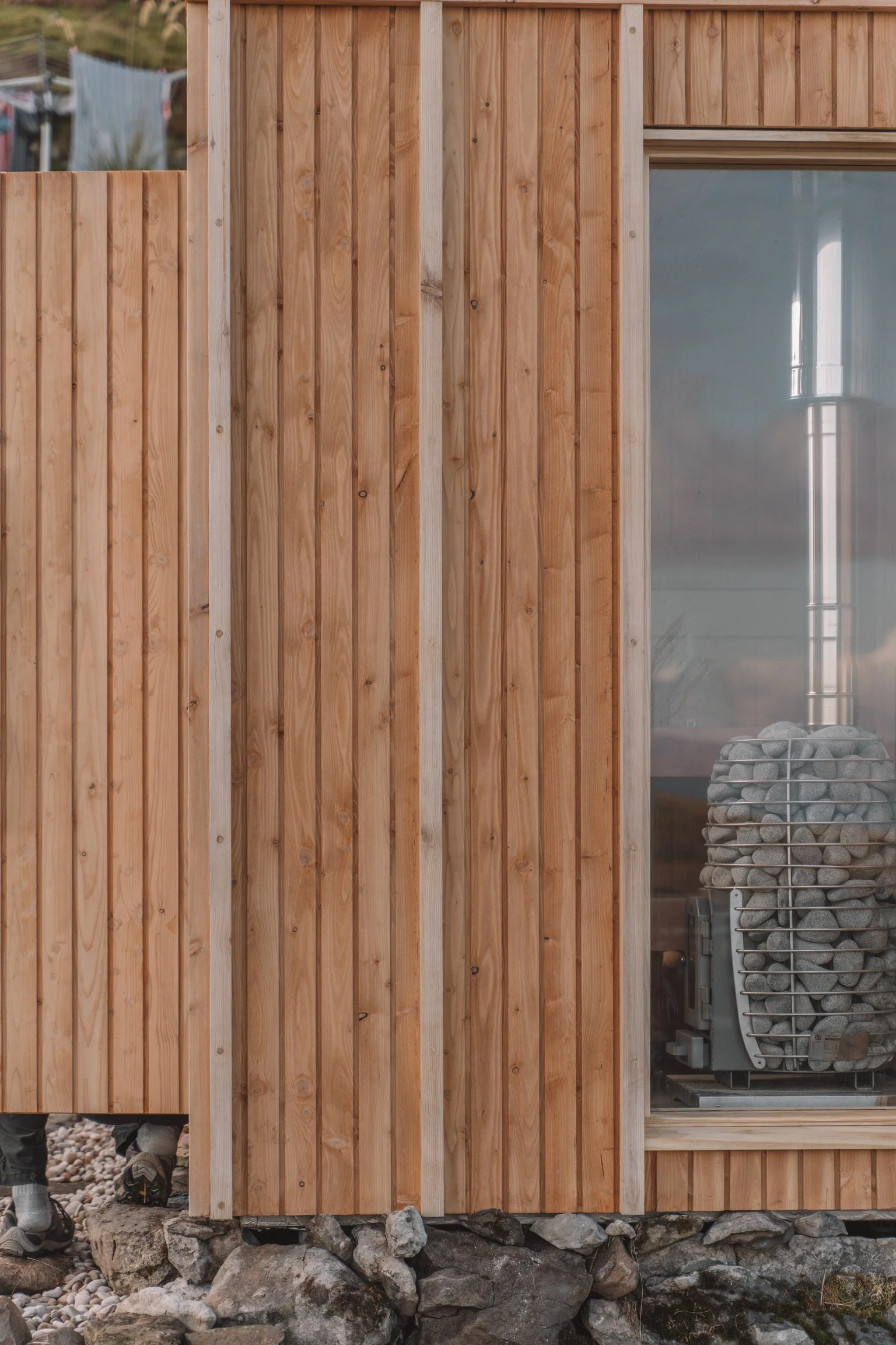 Close-up of a wooden house exterior with vertical wooden planks and a large window, showing rocks and construction equipment inside.