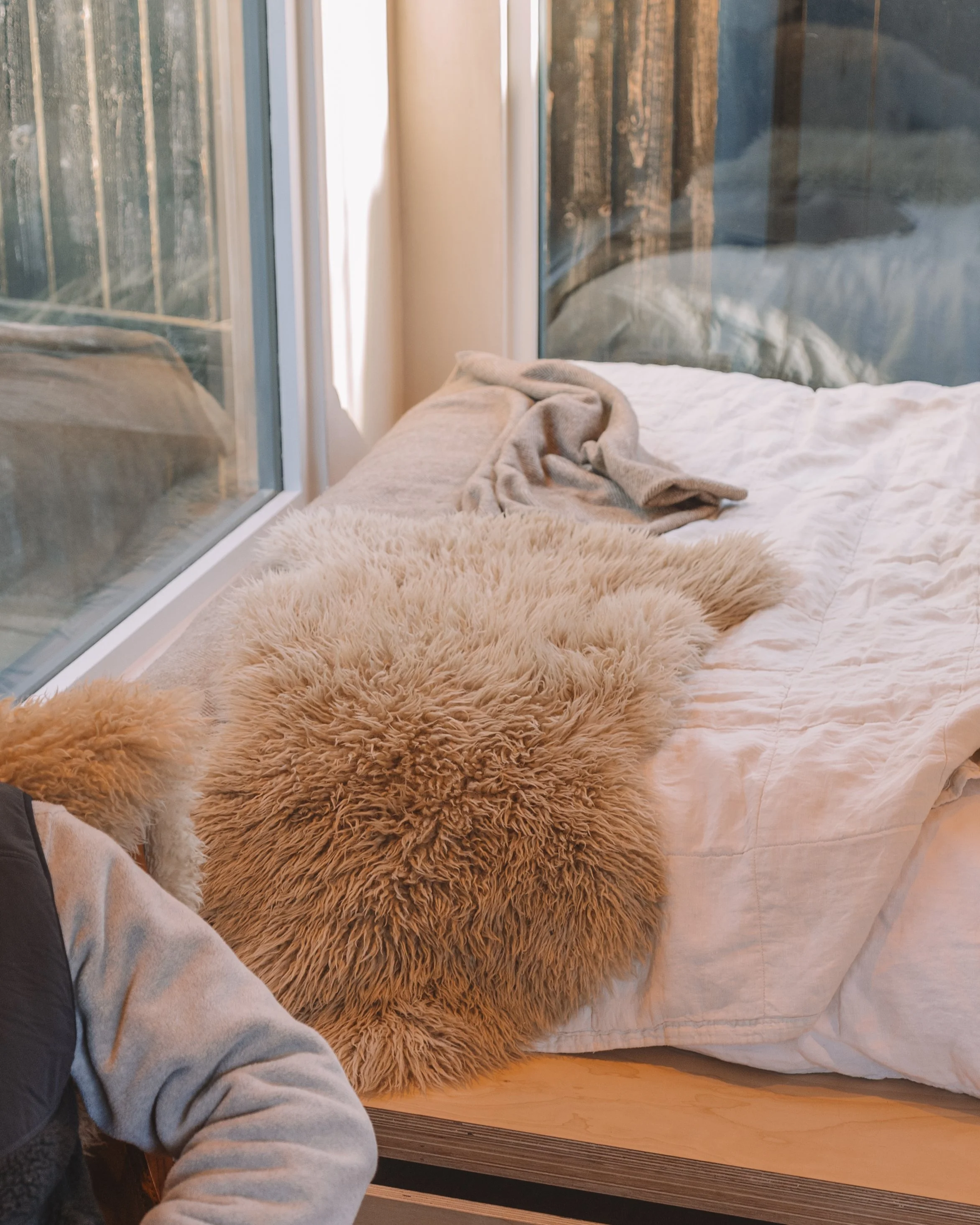 A bed with a white quilt, a beige fluffy rug, and beige blanket near large glass windows showing trees outside.