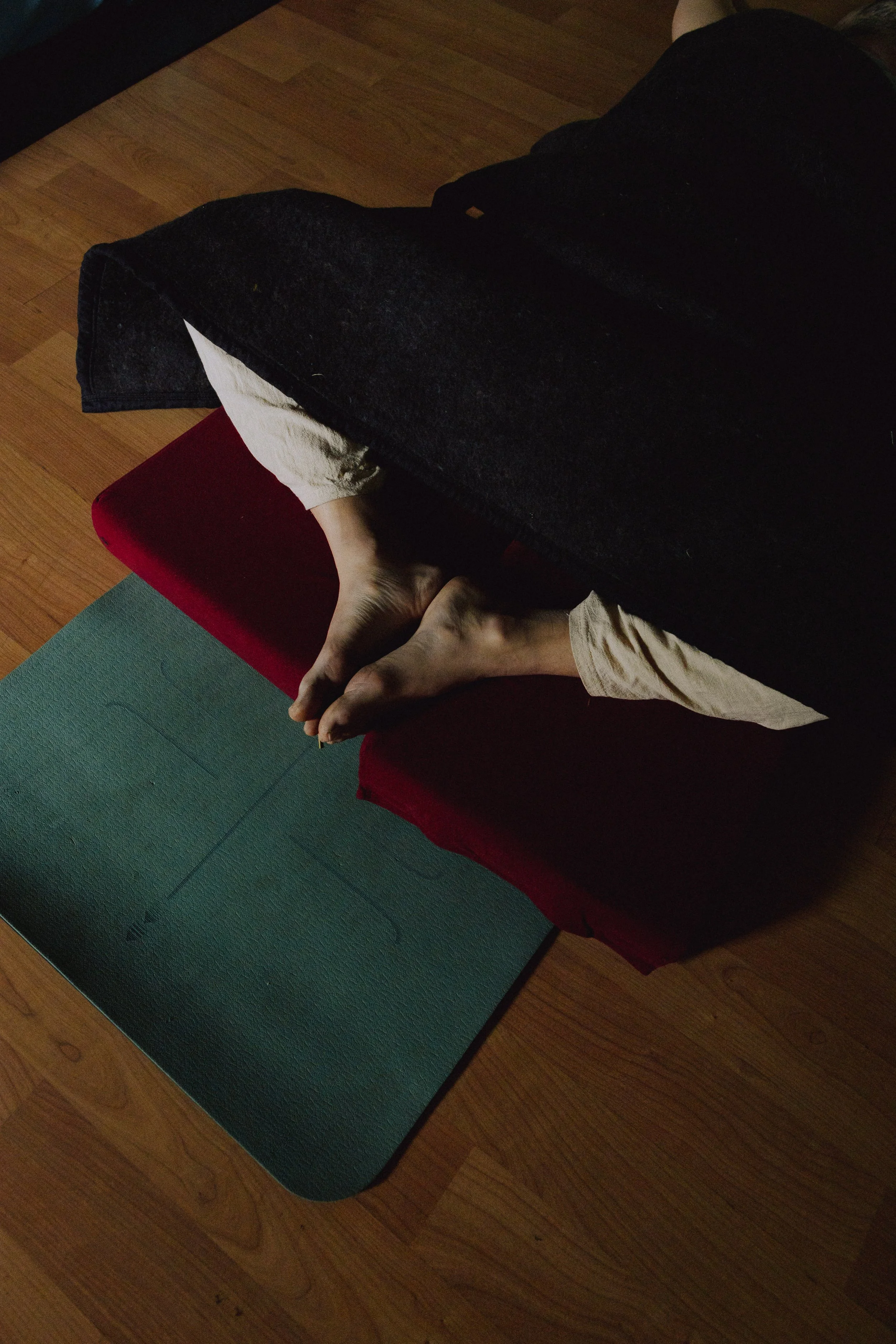 Person sitting on a meditation cushion on a yoga mat with their feet visible, partially covered by a black blanket, in a room with wooden floors.