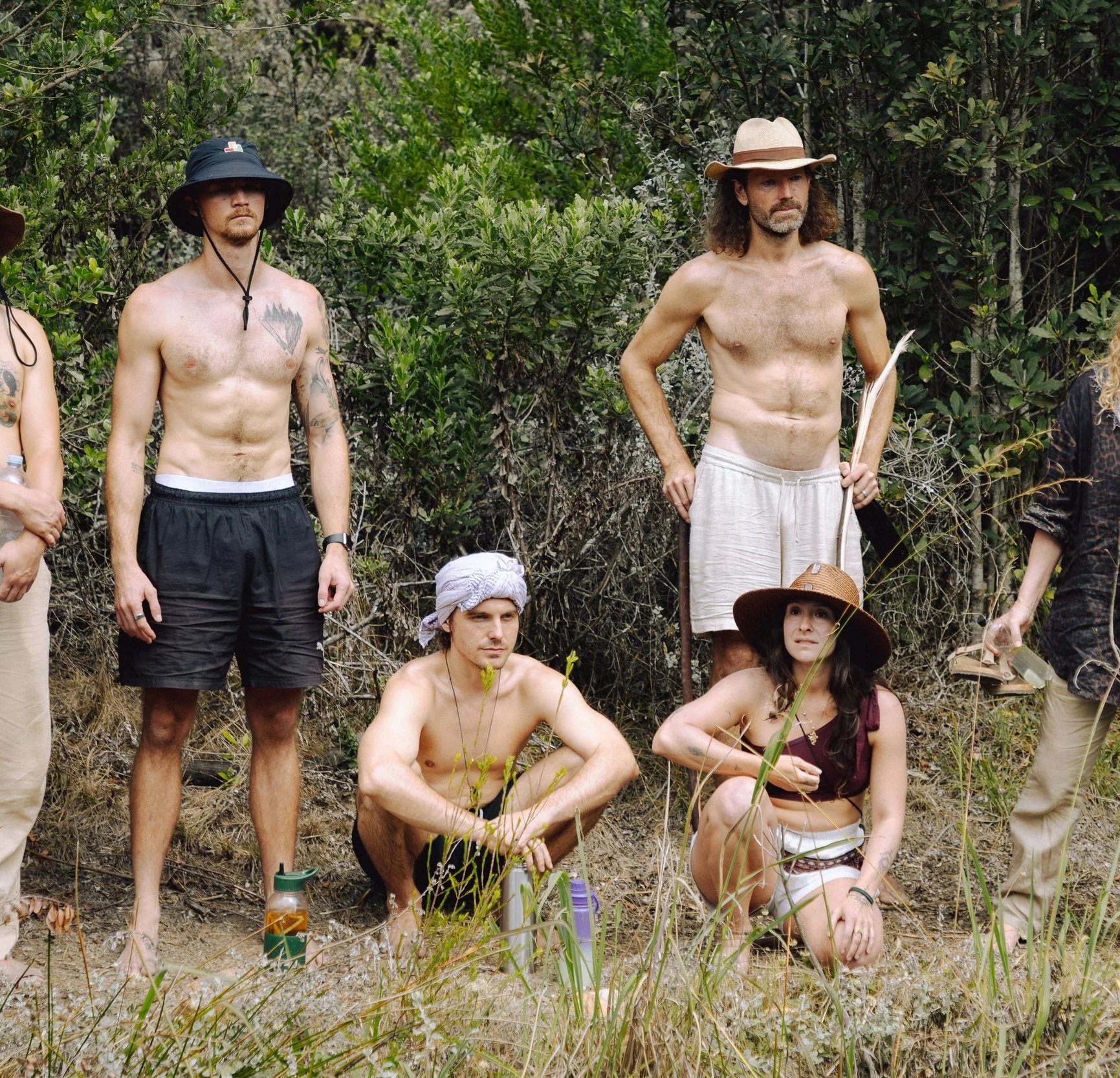 Group of people standing together in a lush, green outdoor setting, some are shirtless, wearing hats, and smiling, suggesting a casual or social gathering in a natural environment.