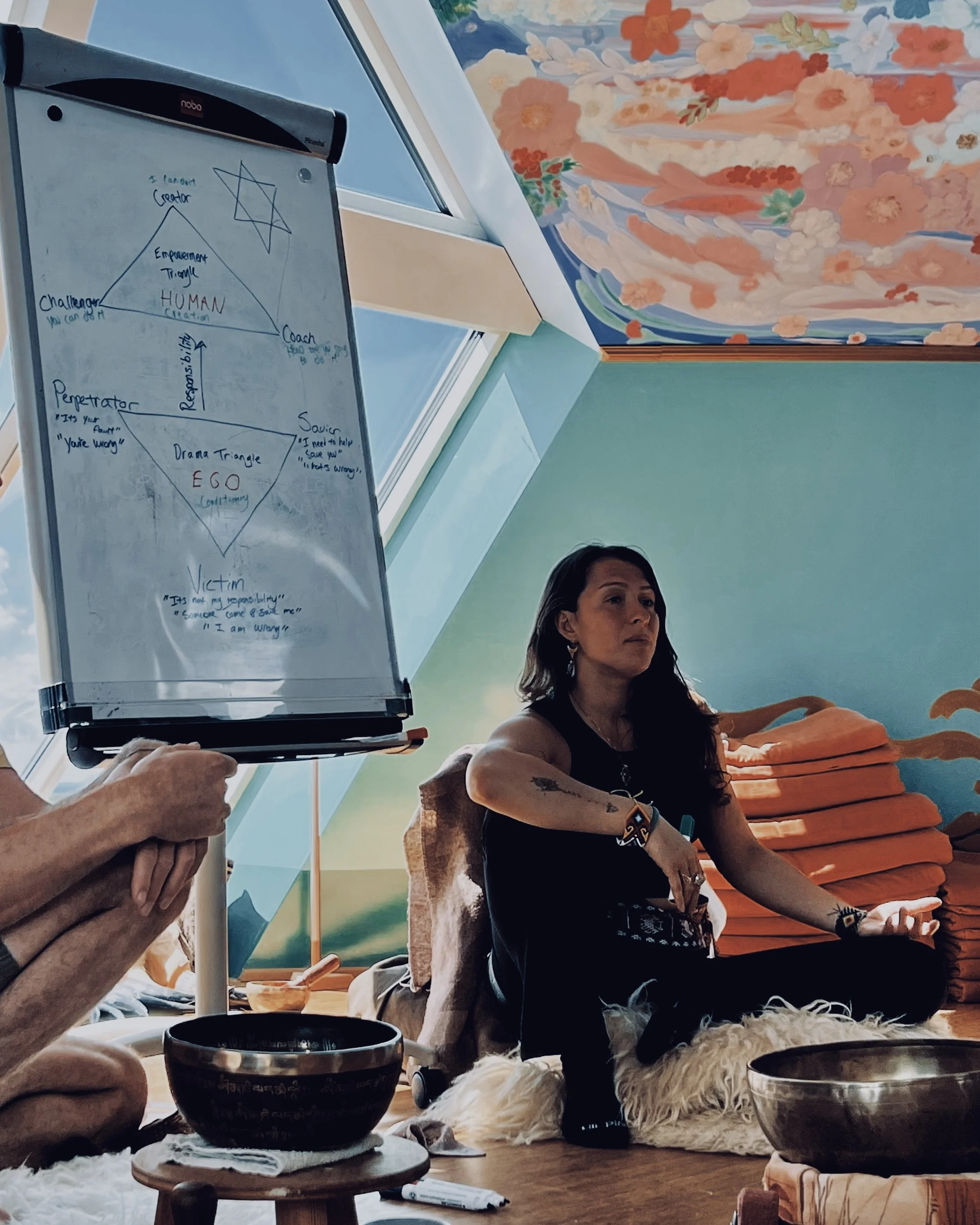 A woman with dark hair, wearing a black spaghetti strap top and gold jewelry, posing inside near a window with city buildings visible outside.