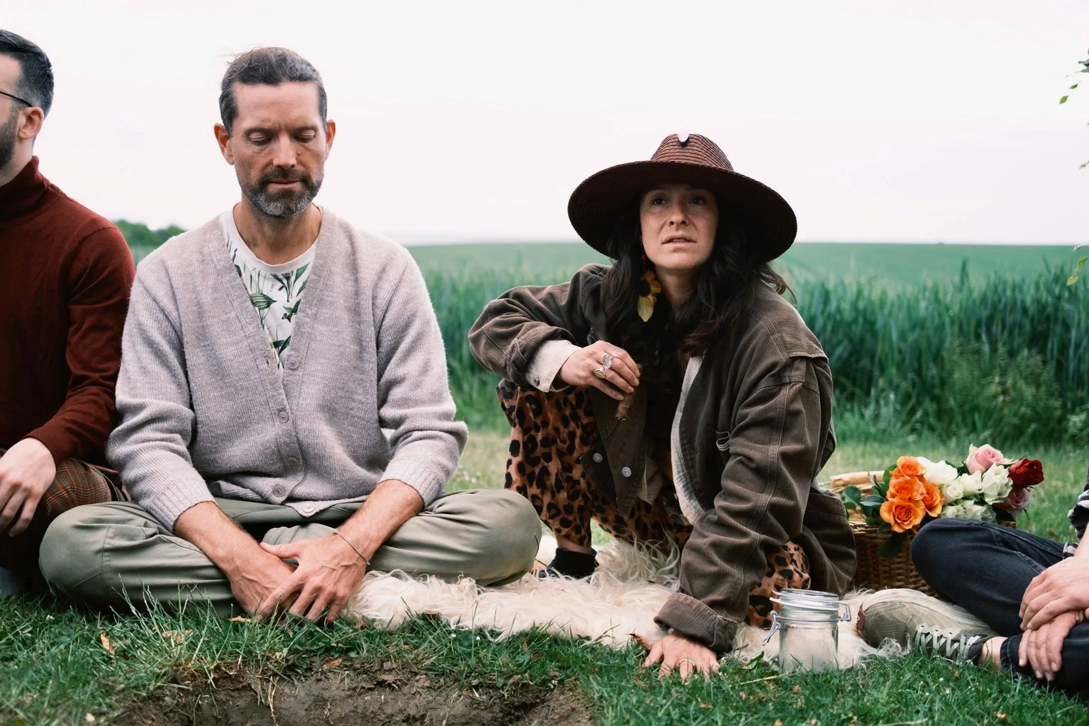 People sitting outdoors on grass, participating in a memorial or ceremony near a grave, with flowers and a basket in the background.