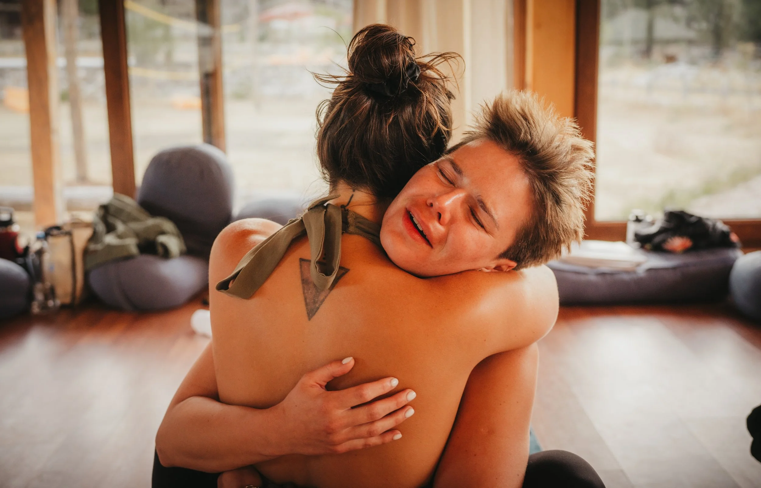 Two people hugging, one with their back to the camera and the other facing the camera, inside a room with large windows and outdoor scenery visible.