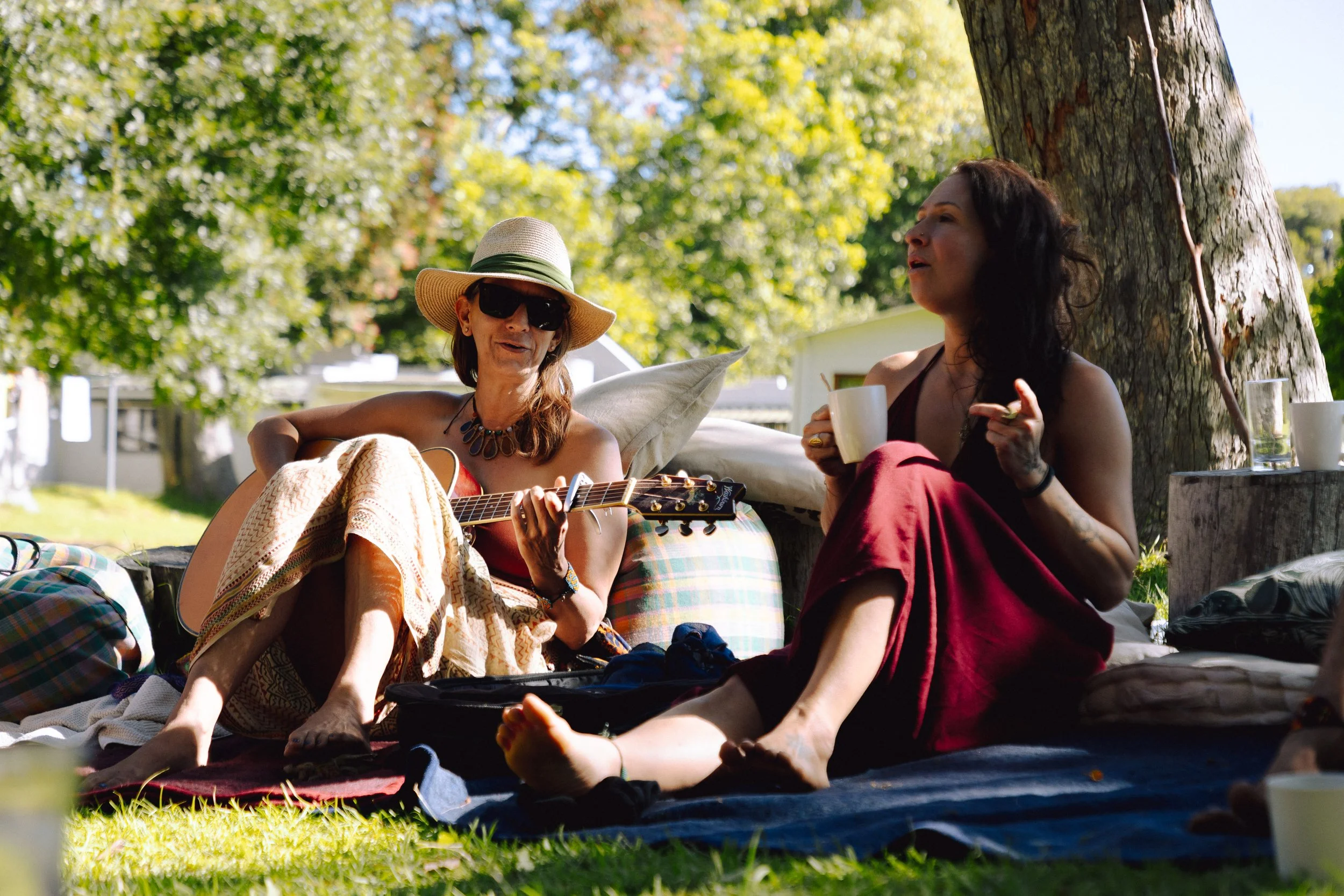 Three women standing outdoors in a grassy clearing surrounded by trees, one holding a stick, engaged in a conversation, with a mountain landscape in the background.