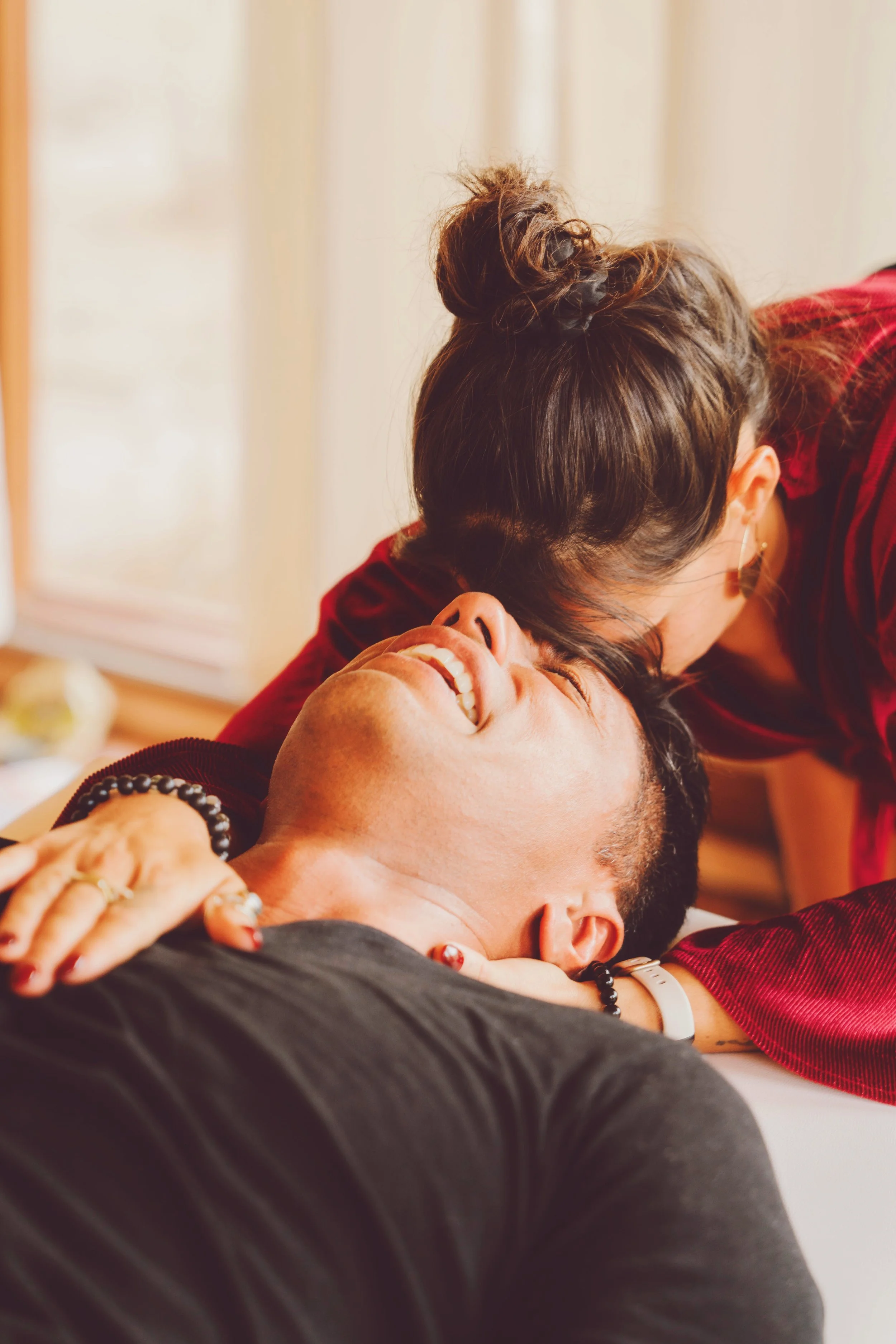 A woman and man share a joyful moment, with the woman kissing the man on the forehead as he lies down, smiling.