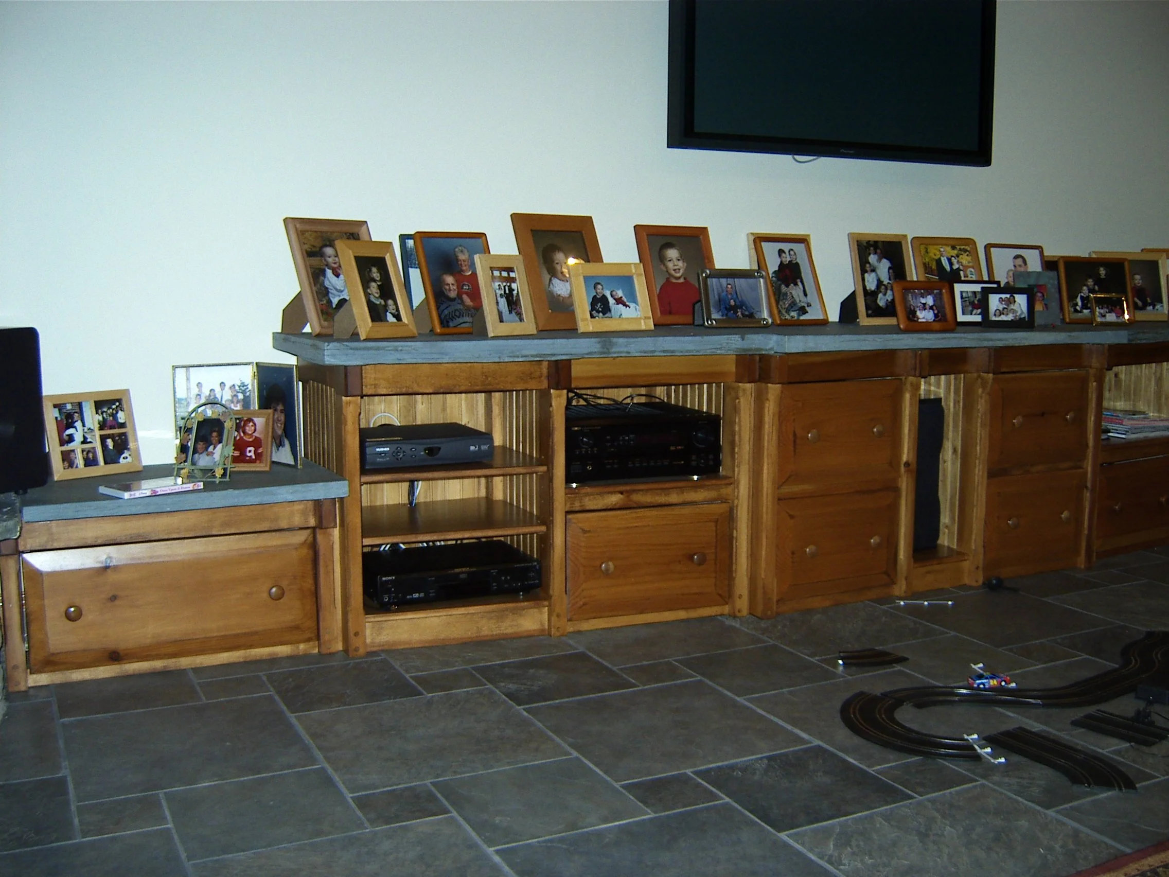 Select Pine custom built entertainment center, with solid blue stone top applied with numerous framed family photographs on top, and a large flat-screen TV mounted on the wall above.