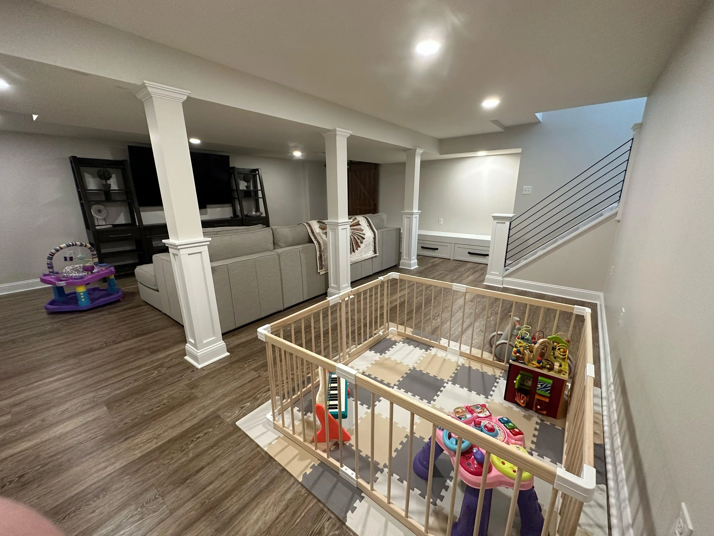 Basement into family room with a baby play area, a large sectional sofa, a television, and staircase with a steel fabricated railing. Columns with molding panels and trimmed details, LED lighting through out new interlocking floor commercial grade