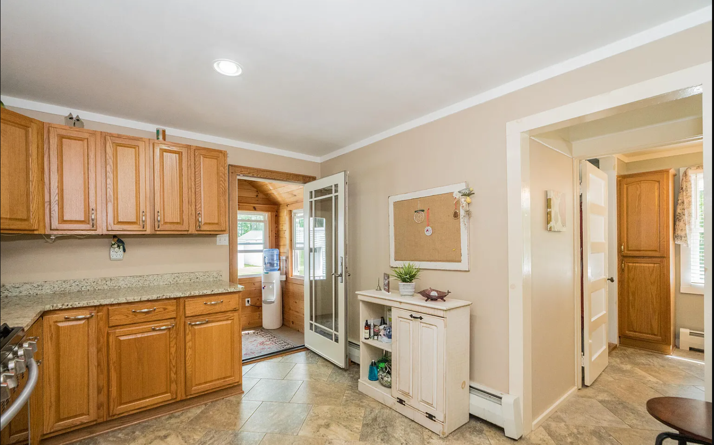 Kitchen with oak cabinets, granite countertops, open door leading to a small room pine tongue and grove with water dispenser, beige walls, and decor including a plant and small ornaments.