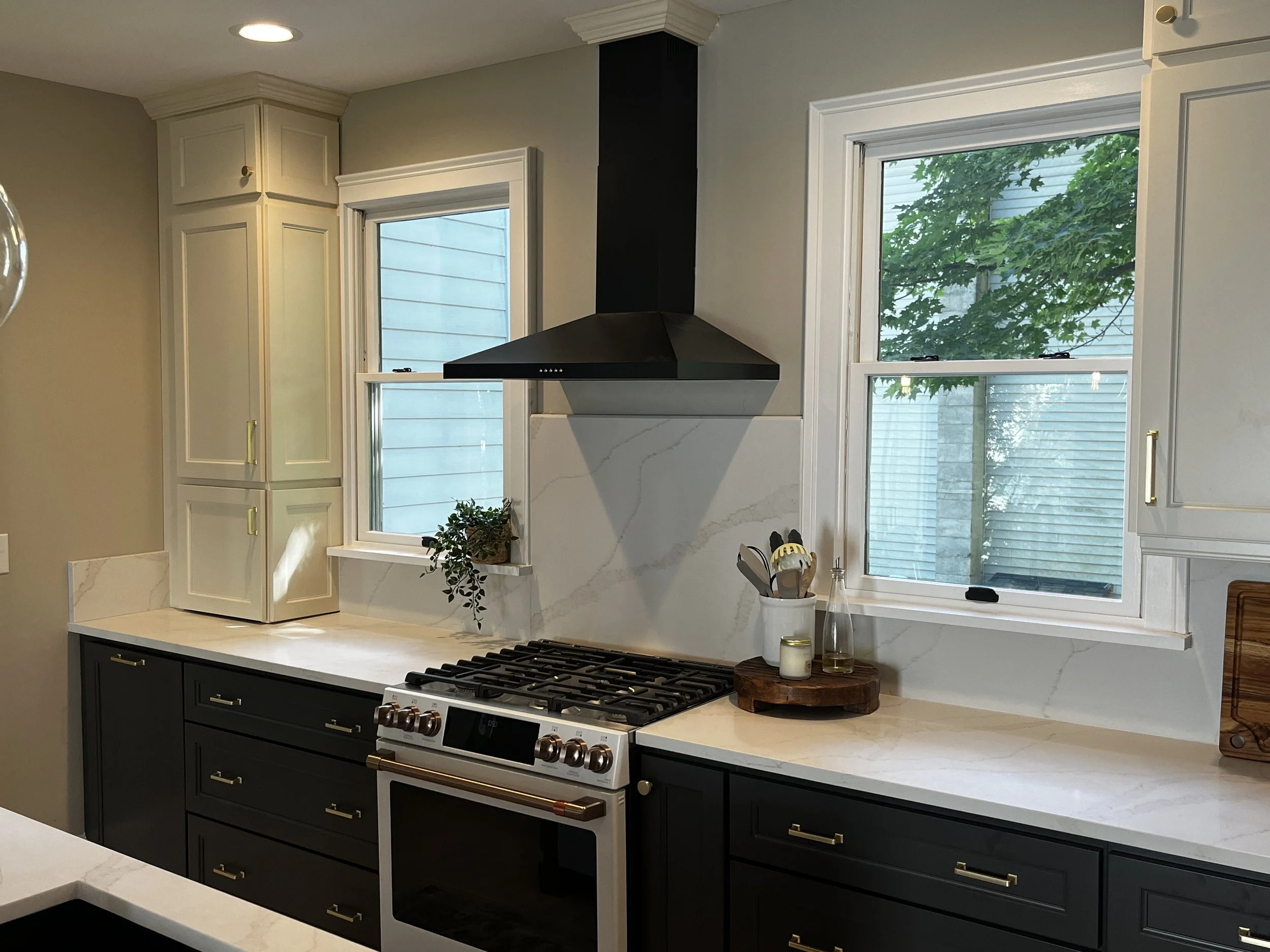 newly remodeled kitchen with white quartz countertop , forest green base cabinets, pearl color upper cabinets with garage on the left, a gas range, range hood, black coated exhaust hood and two new double hung windows with window casing