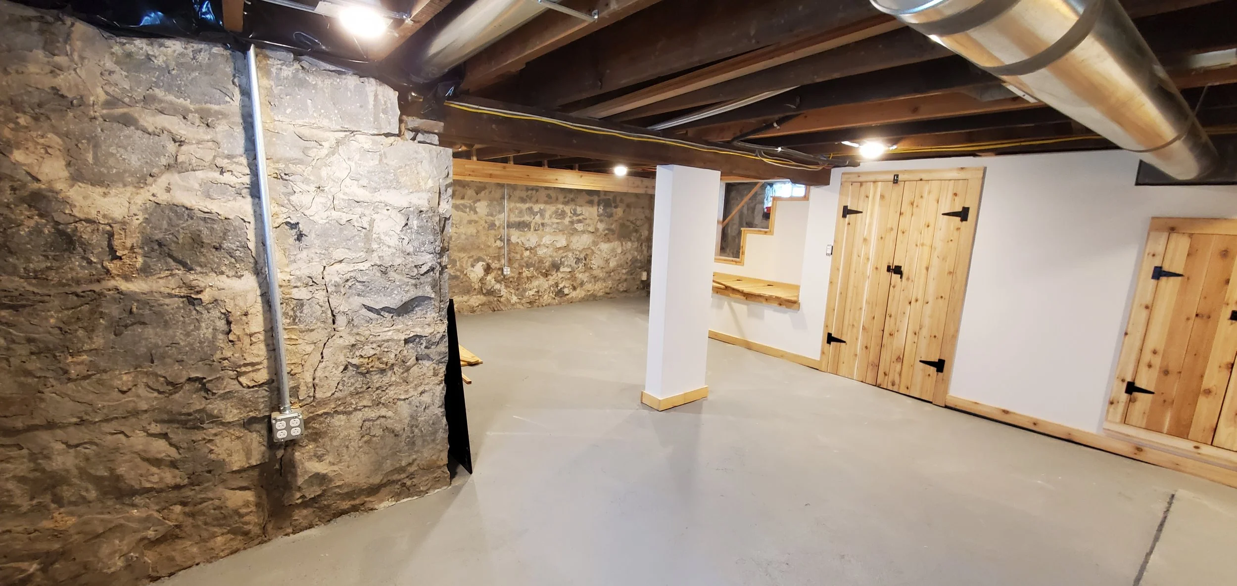 Unfinished basement with stone wall, exposed wooden ceiling beams, concrete floor, and wooden doors.