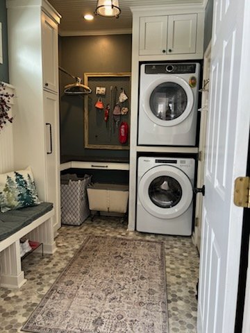 Custom built laundry and mudroom with stacked washer and dryer, cabinetry, a bench with a pillow, a laundry hamper, and a wall with hooks and hanging items.