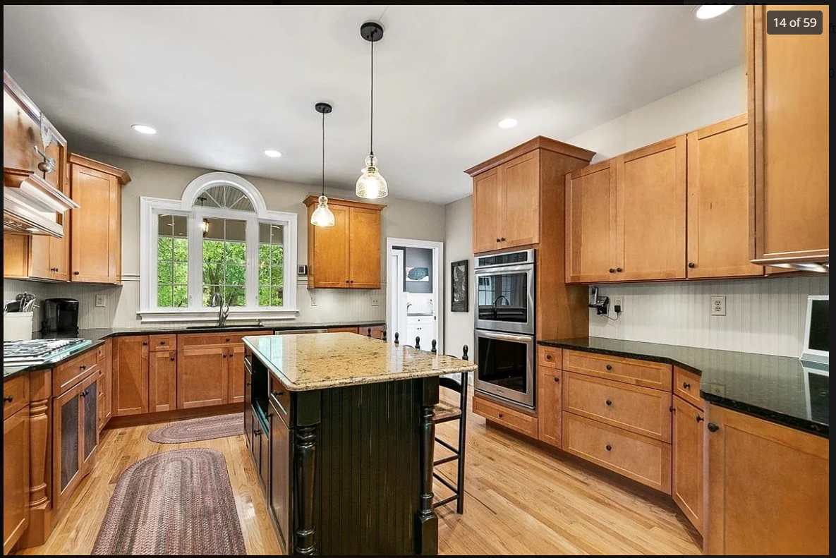 A spacious kitchen with wooden cabinets, a central island with a granite countertop, and a large window overlooking green trees. The kitchen has pendant lights and built-in modern appliances, including double ovens and a stovetop.