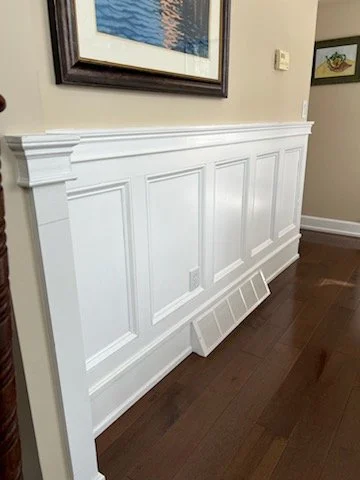 White wooden wainscoting with decorative panels and a vent cover on the floor in a room with hardwood floors and framed artwork on the wall.