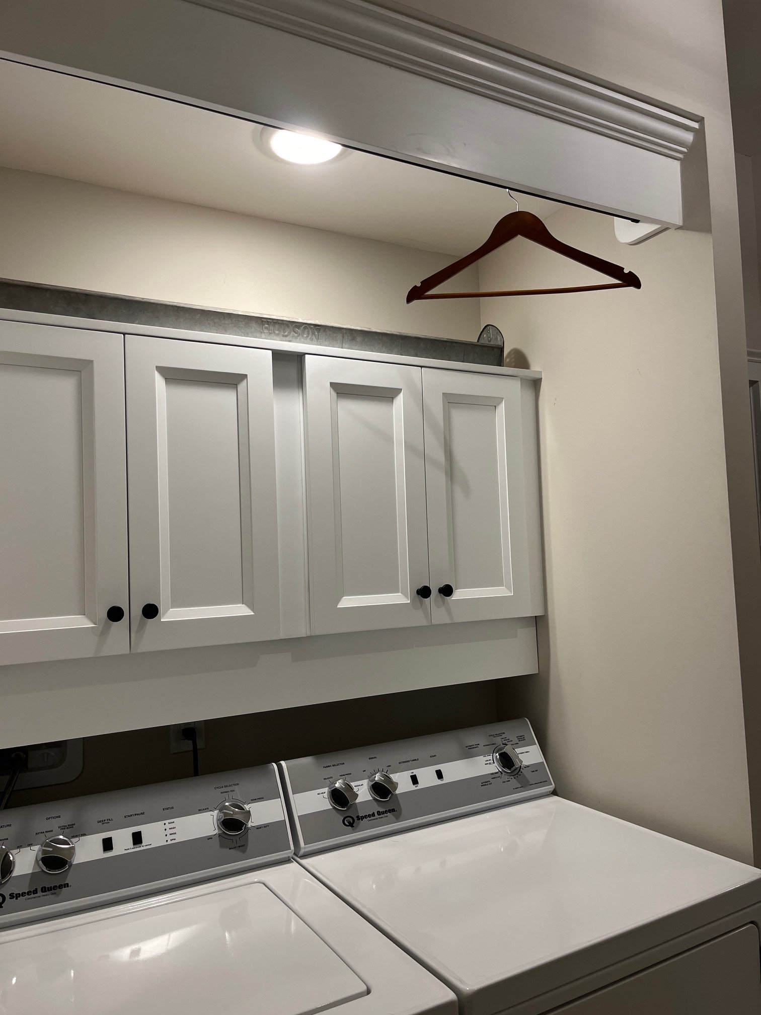 A laundry room with two white washing machines, white cabinets above, a metal rod with a wooden hanger, and a ceiling light.