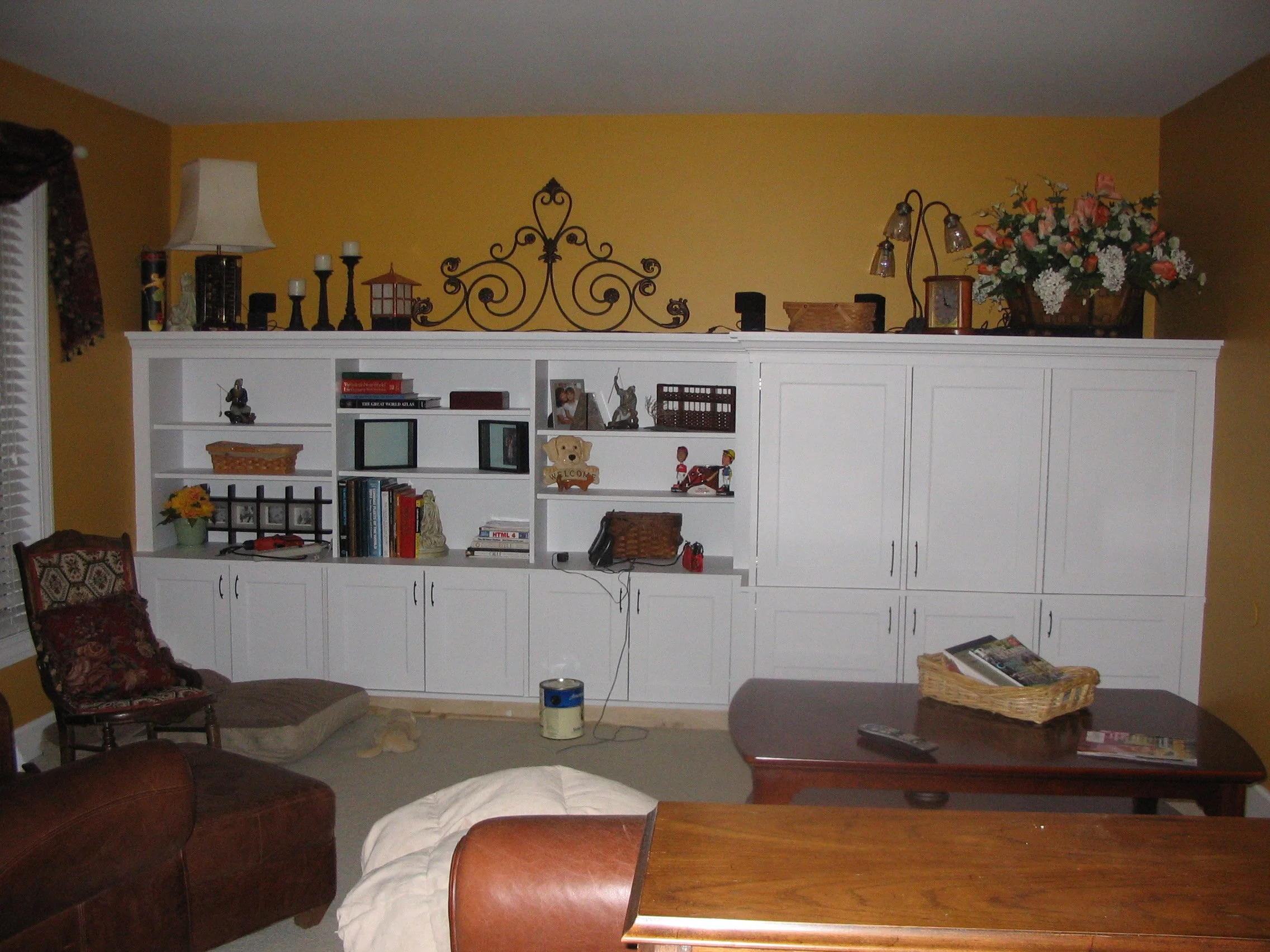 Living room with white built-in shelves, yellow walls, and hardwood table, decorated with books, pictures, a lamp, and a large flower arrangement.