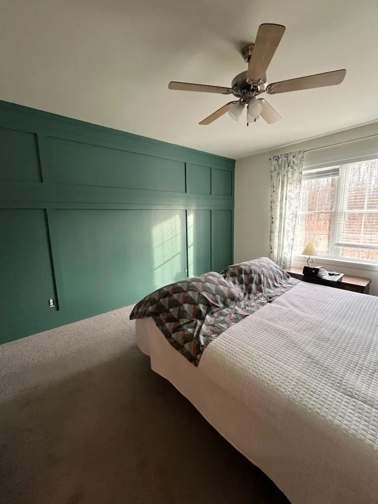 Bedroom with green accent wall, bed with patterned pillows and white quilt, window with floral curtains, ceiling fan, nightstand with lamp and clock, beige carpet.