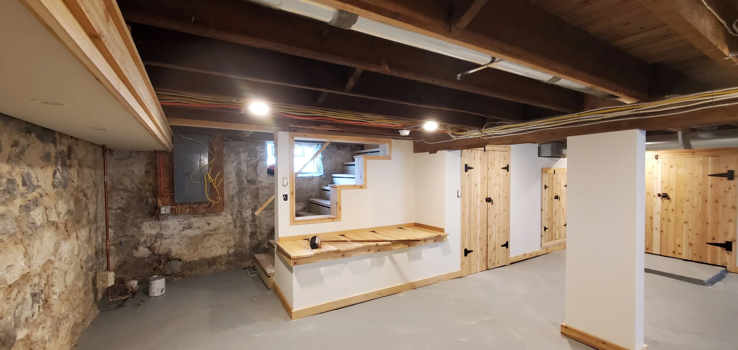 Basement under renovation with exposed wooden beams, unfinished stone walls, and new wooden doors and trim. There are stairs and small windows, with construction tools and wires visible.