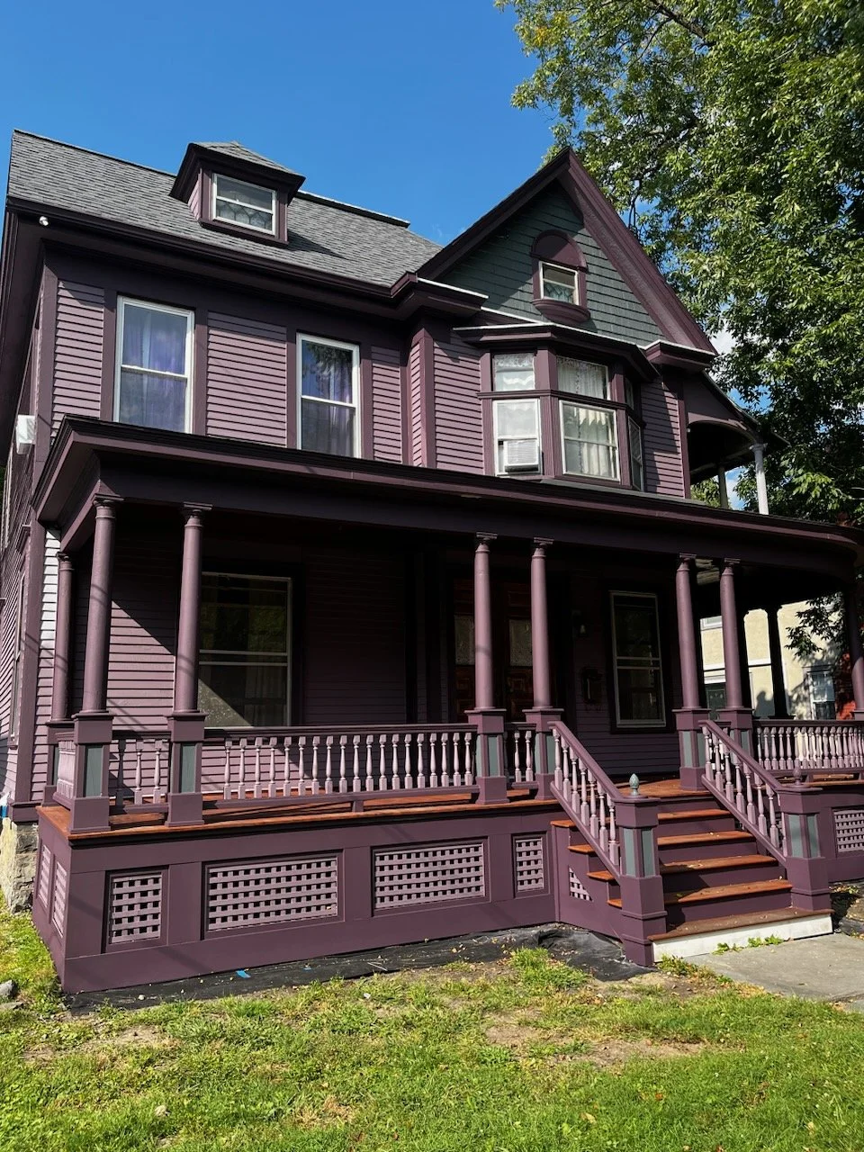 A three-story Victorian-style house painted in purple with a front porch supported by multiple purple columns, wooden stairs leading to the porch, and several windows, including a bay window on the second floor. There are trees and a blue sky in the 