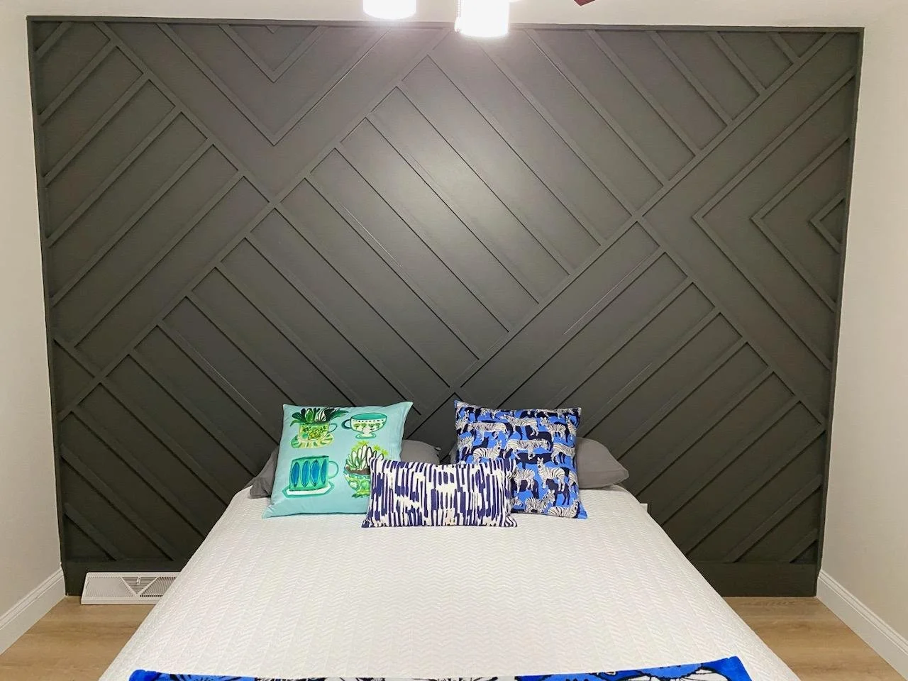 A bed with a white bedspread against a black textured wooden headboard with diagonal patterns. There are four decorative pillows on the bed with various blue, green, and patterned designs.