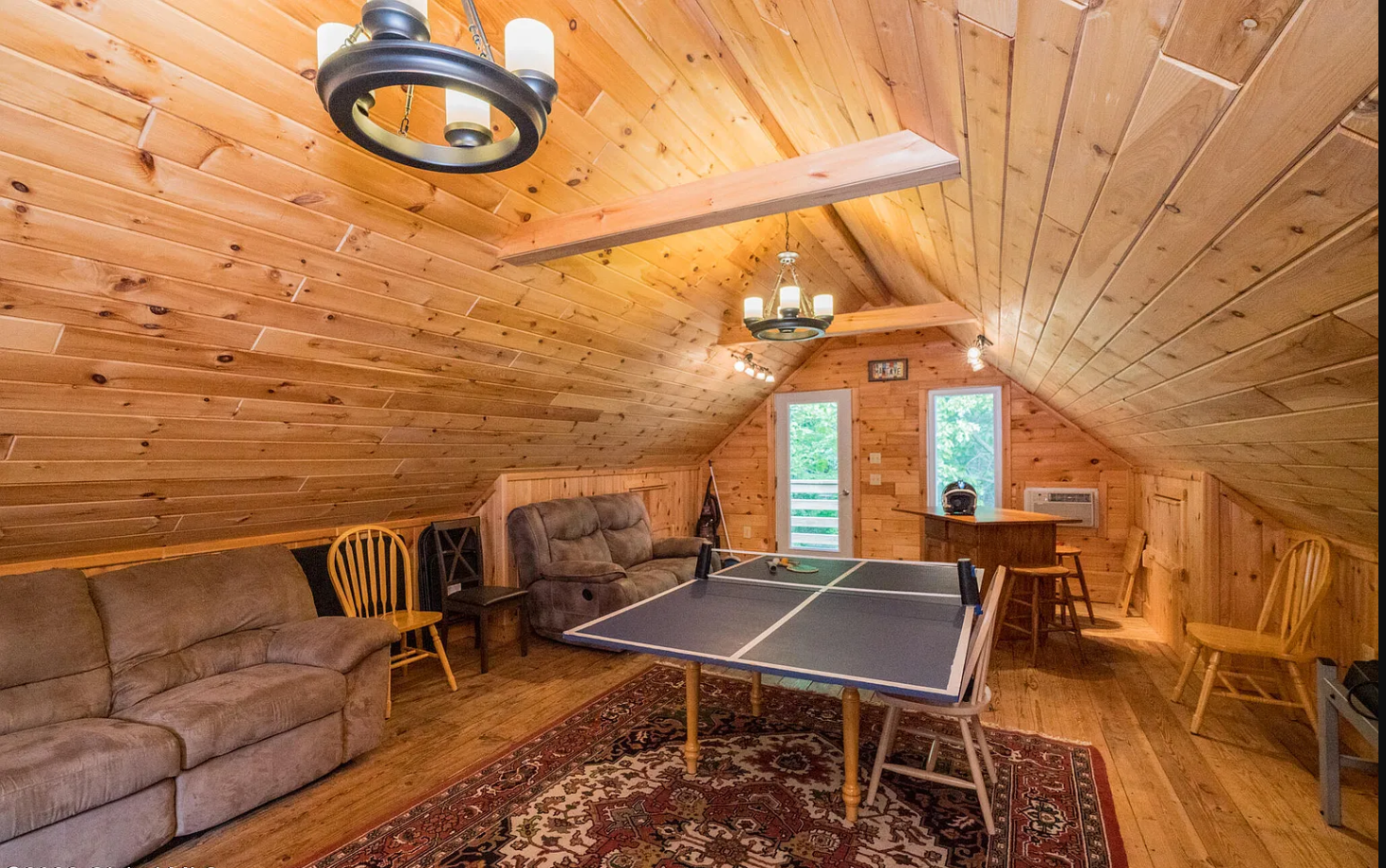 Loft game room with wood-paneled walls and ceiling, featuring a ping pong table, sofas, chairs, a bar counter with a stool, and windows with views of greenery.