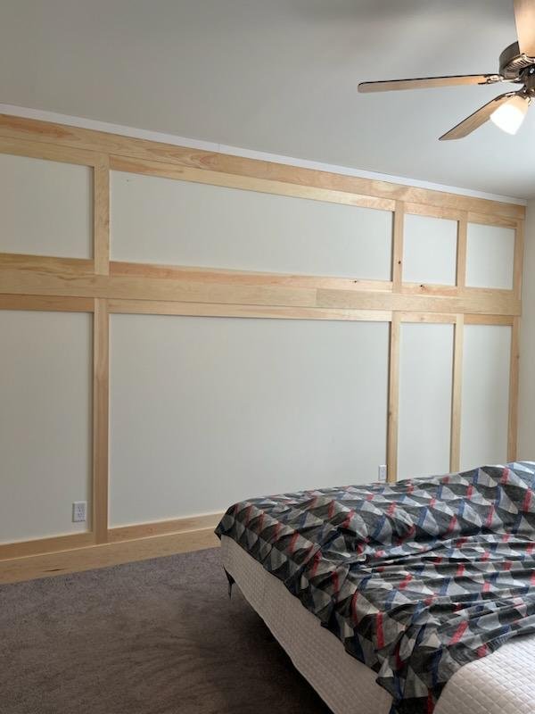 Interior of a bedroom with a partially finished wooden wall and a ceiling fan. The bed has a patterned comforter.