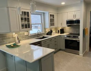 Modern kitchen with white cabinets, gray countertops, and stainless steel appliances, including a stove and microwave. There is a window above the sink, and the backsplash is made of light gray tiles.