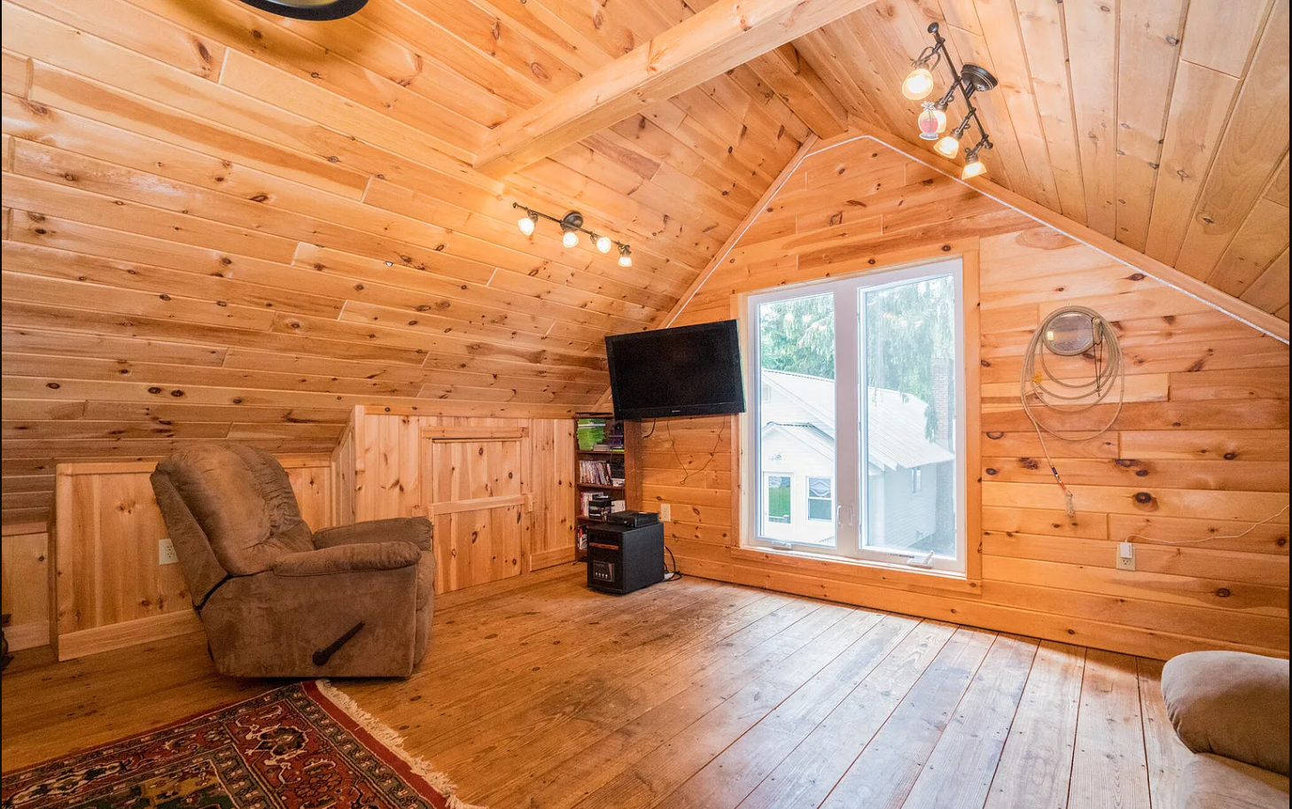 A cozy attic room with wooden paneling on the walls and ceiling, a sliding glass door leading outside, a wall-mounted TV, a small bookshelf with books, a brown recliner chair, a side table, and exposed electrical wiring.