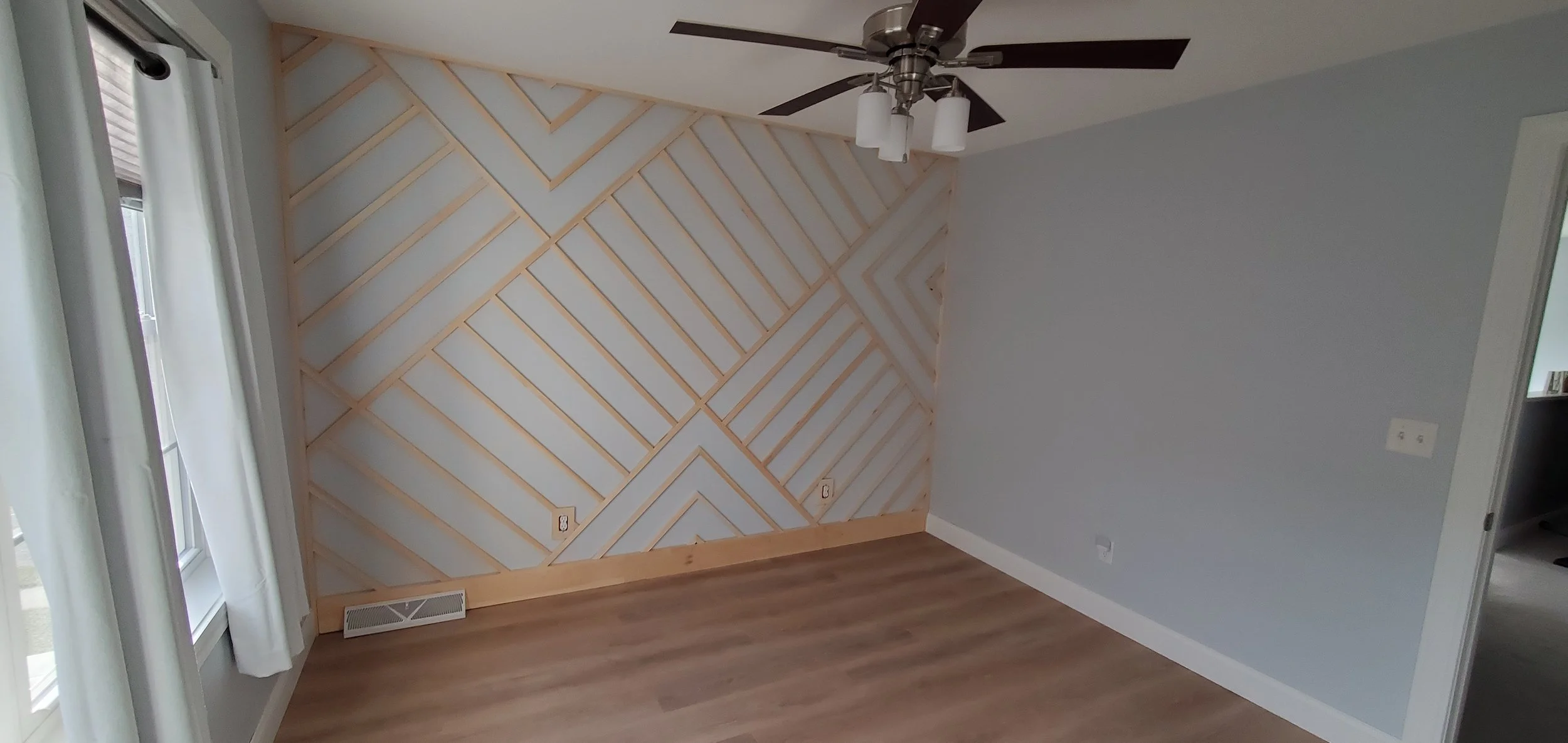 Empty room with light gray walls, wooden flooring, a corner wall with a geometric wooden slat design, a ceiling fan with dark blades and three light fixtures, a window with white curtains, and a door leading to another room.
