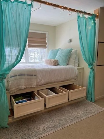 A cozy bedroom with a bed covered in white bedding and Oversized pillows. The bed is surrounded by turquoise curtains tied back, with a window in the background. The bed frame has built-in storage drawers underneath.