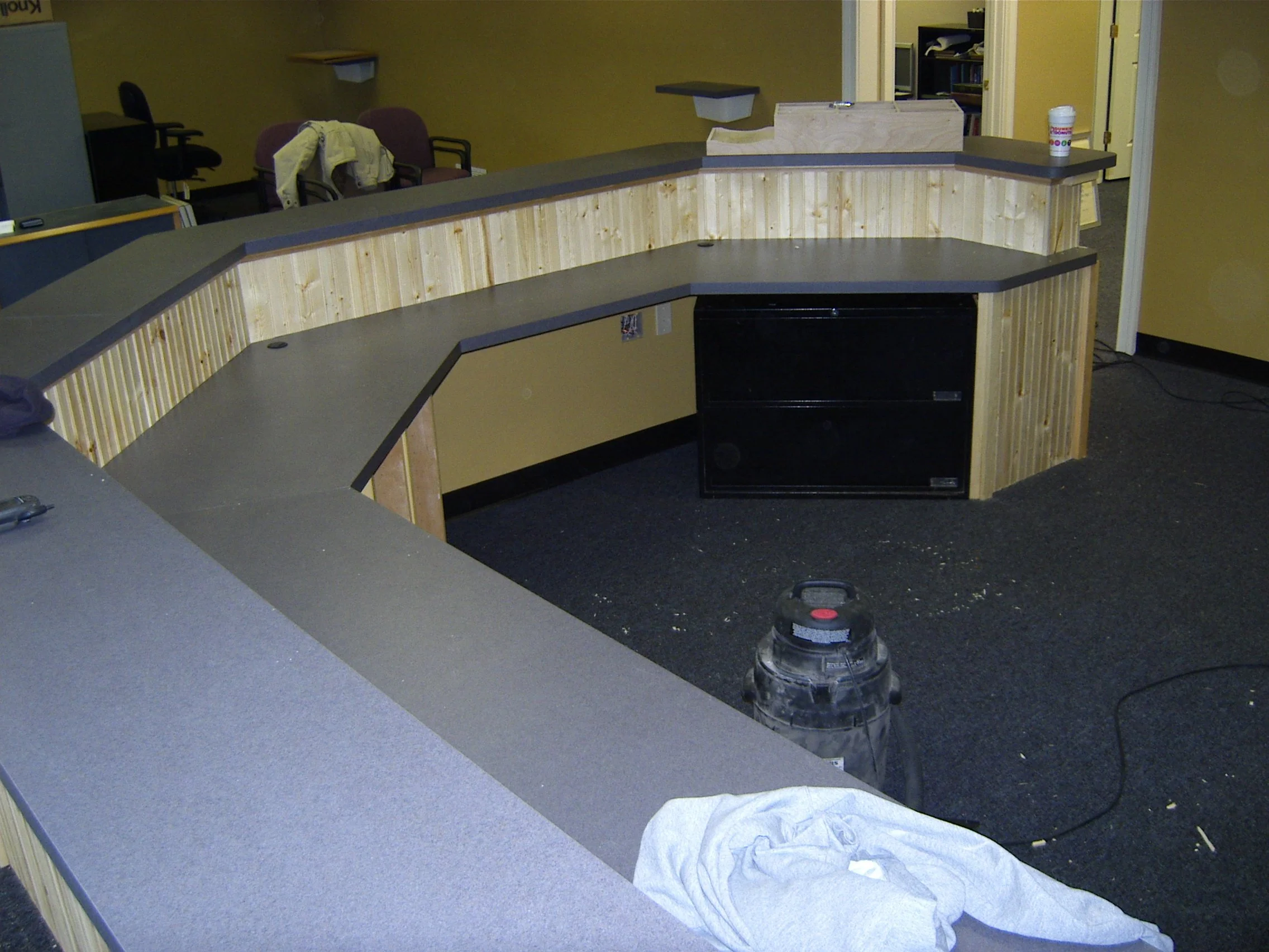 An empty office or reception area with a curved wooden counter, a black filing cabinet underneath, a vacuum cleaner on the dark carpet, and some chairs and desks in the background.